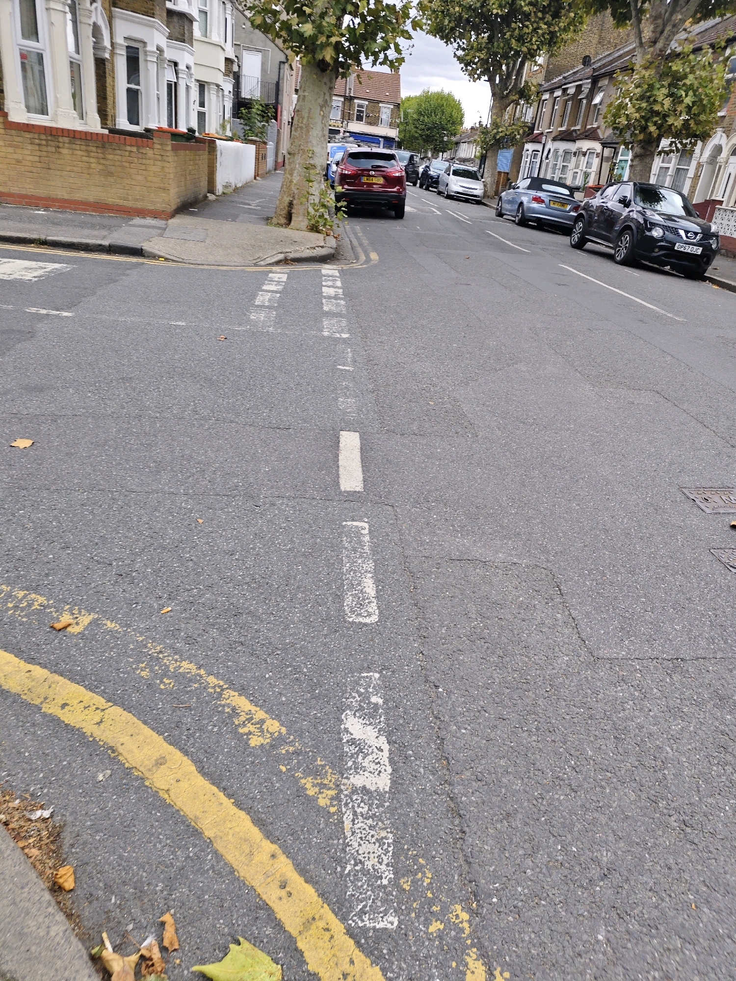 A street scene in London, United Kingdom, is depicted in the image. The viewpoint is at street level, looking down the road. The road surface is asphalt, with white and yellow painted lines marking lanes and parking restrictions. Several parked cars are visible along the side of the road, including a black Nissan Juke, a silver convertible, and a red SUV. Houses line the street on both sides, characterized by their brickwork and traditional architectural features. Trees with green foliage are scattered along the pavement. The sky is overcast, suggesting a cloudy day. There are no people visible in the frame.