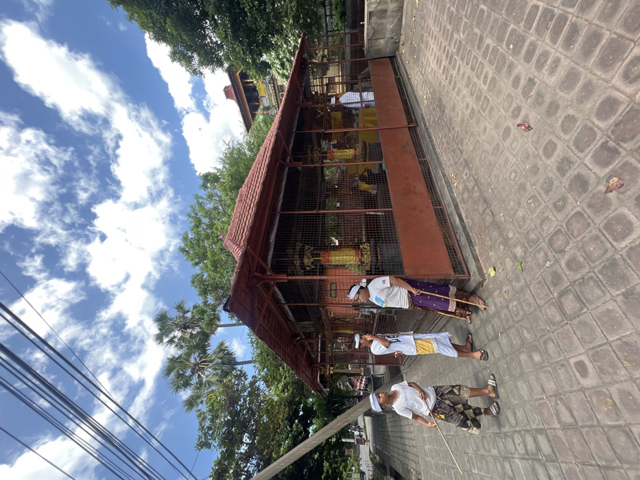 This image depicts an outdoor scene in Banyu Poh, Indonesia, under a bright blue sky with scattered white clouds. In the foreground, three males are present on a paved area with interlocking square tiles. All three are wearing white head coverings, white t-shirts, and patterned sarong-like lower garments (kamen); two are wearing flip-flops. The leftmost individual wears a patterned brown and white sarong, a white t-shirt, and holds a thin wooden stick in his left hand, looking towards the center. The middle individual wears a white and yellow patterned sarong, a white t-shirt, and holds his right hand near his mouth, facing the rightmost person. The rightmost individual wears a dark purple patterned sarong, a white t-shirt with a graphic design, and leans against a building while holding a thin wooden stick with both hands.
The primary subject is a single-story rectangular building with a red tiled roof and a brown wooden or metal frame, enclosed by metal mesh or bars. The lower section of the building's walls is solid brown. Inside the building, several golden-colored structures, possibly altars or offerings, are visible. Another person in similar attire is partially visible inside the building on the far right.
To the left of the scene, a utility pole slants upwards and left, with multiple overhead lines crossing the upper left quadrant of the sky. A road or street is visible behind the pole, with distant vehicles or structures obscured by foliage. The background features lush green trees, including several tall palm trees with visible fronds. To the far right, behind the enclosed building, another multi-tiered structure with a red roof and yellow details is partially visible, indicating a larger temple complex. The lighting suggests a sunny day.