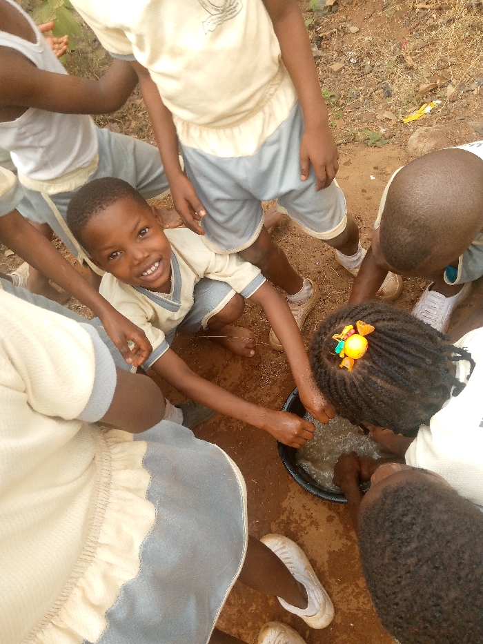 The image depicts an overhead view of six children gathered on reddish-brown dirt ground around a black plastic basin containing murky water in Ukwu-Abwa, Nigeria. A male child, centrally positioned and kneeling, wears a light yellow shirt with a blue trim and matching shorts, is barefoot, and smiles directly at the camera with his right arm extended towards the basin. To his right, another child with braided hair, adorned with a yellow and green hair clip, leans over the basin with both hands immersed. A third child, partially visible from behind, also has hands in the basin. Other children, partially visible around the periphery, wear similar light yellow/cream tops and light blue shorts, with one child in the upper left wearing a white tank top. Footwear varies from bare feet to light brown shoes and white slip-on shoes. The ground is uneven, scattered with small stones and minor debris. Green foliage is visible in the upper left background. The scene captures a group engaged in an outdoor activity involving water.