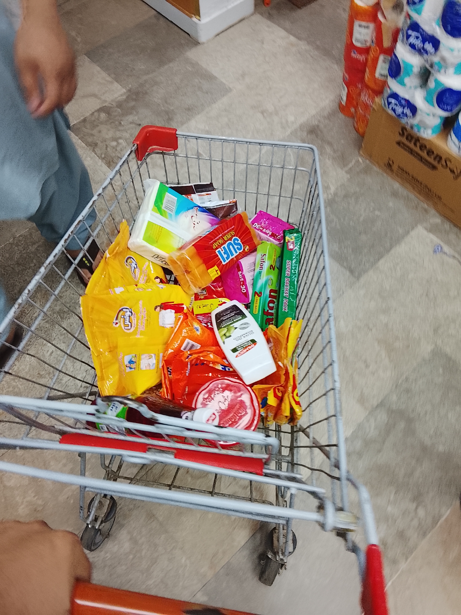 The image shows a shopping cart filled with various grocery items in what appears to be a store or supermarket. The items include multiple packs of detergent powder, specifically 'Ujala Supreme', along with bars of soap, 'Sufi' and 'Safon'. Other items such as shampoo and possibly other household products are also visible. The shopping cart is being pushed by someone in a store, as indicated by the flooring and surrounding items. The scene is likely taking place in the city of Gulgasht Colony, Pakistan, given that the detergent brand and products are commonly available in that region.