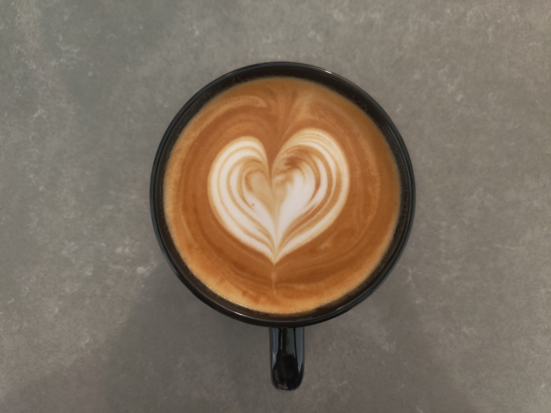 A top-down, medium shot captures a black ceramic mug filled with latte art, resembling a heart shape, resting on a speckled grey surface. The mug handle is visible at the bottom of the frame. The background is a plain, textured grey surface. The lighting is consistent, casting no harsh shadows, suggesting an indoor setting. The image provides no information about the city of Kelowna, Canada.