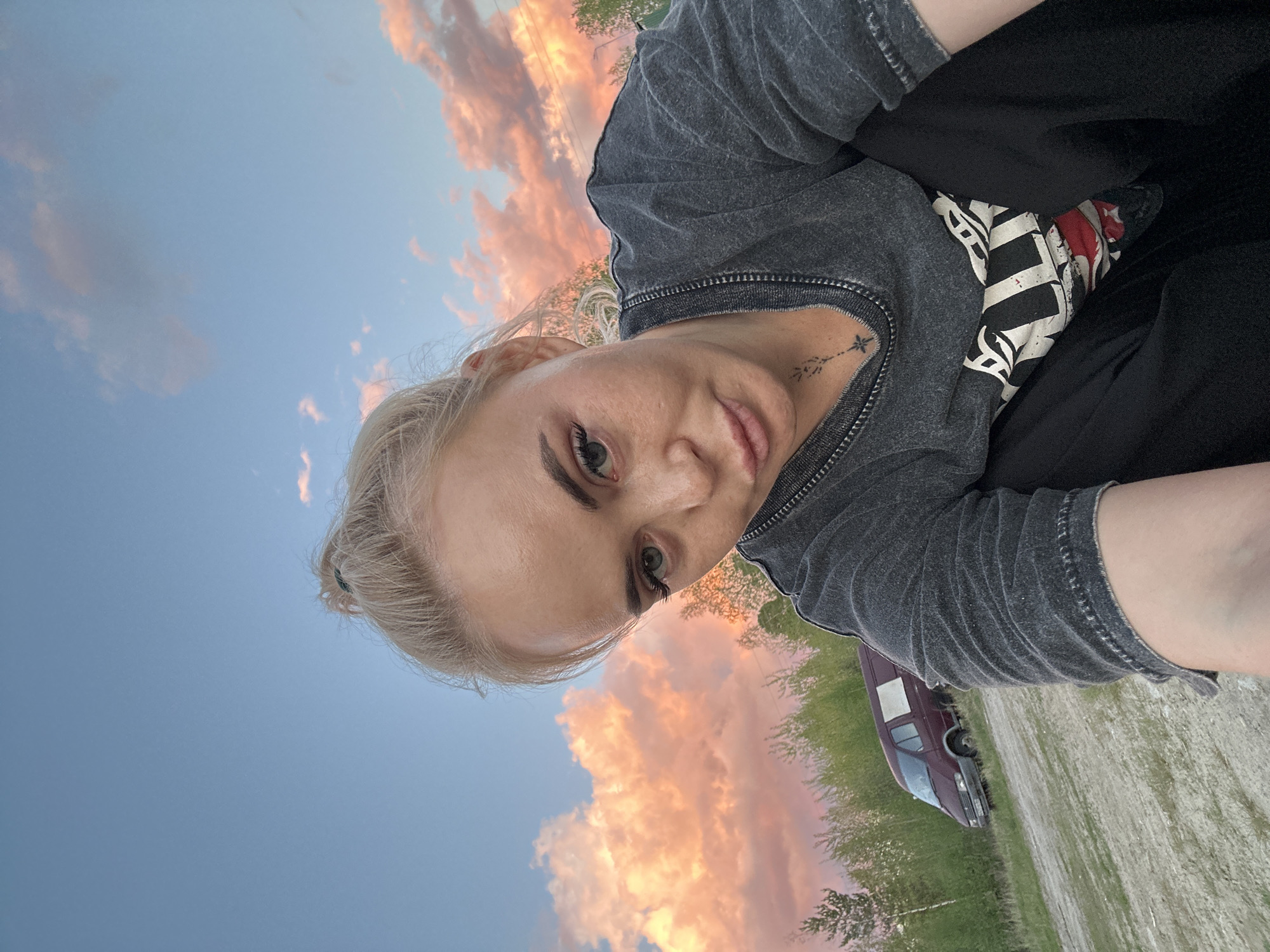 The image, correctly rotated, captures a young woman taking a selfie outdoors against a picturesque sunset sky. The woman, with light blonde hair pulled back into a bun, occupies the left and central foreground, looking directly towards the camera with a calm, neutral expression. She has fair skin, light-colored eyes, and well-defined eyebrows. Her lips are slightly parted.
She is wearing a dark gray, possibly distressed or acid-wash, short-sleeved t-shirt with a ribbed neckline and cuffs. A small, delicate black tattoo is visible on the right side of her neck, resembling a stylized arrow or stem topped with a star or floral design, accompanied by small, illegible characters below. A black, white, and red patterned piece of fabric or clothing is partially visible near her right hip, and she appears to be wearing black pants or shorts.
The background is dominated by a vast sky filled with beautifully colored clouds, displaying shades of pink, orange, and red, suggesting either sunrise or sunset, with the latter being more probable due to the warm, low light illuminating the clouds. The sky transitions to a softer blue overhead. In the mid-ground, green foliage and trees are visible, and to the lower right, a dark red or maroon van or large car is parked on a dirt or gravel path or road. The setting appears to be a natural, open outdoor area, consistent with the provided location context of Высокий, Russia. No discernible English text is visible apart from the intricate tattoo.