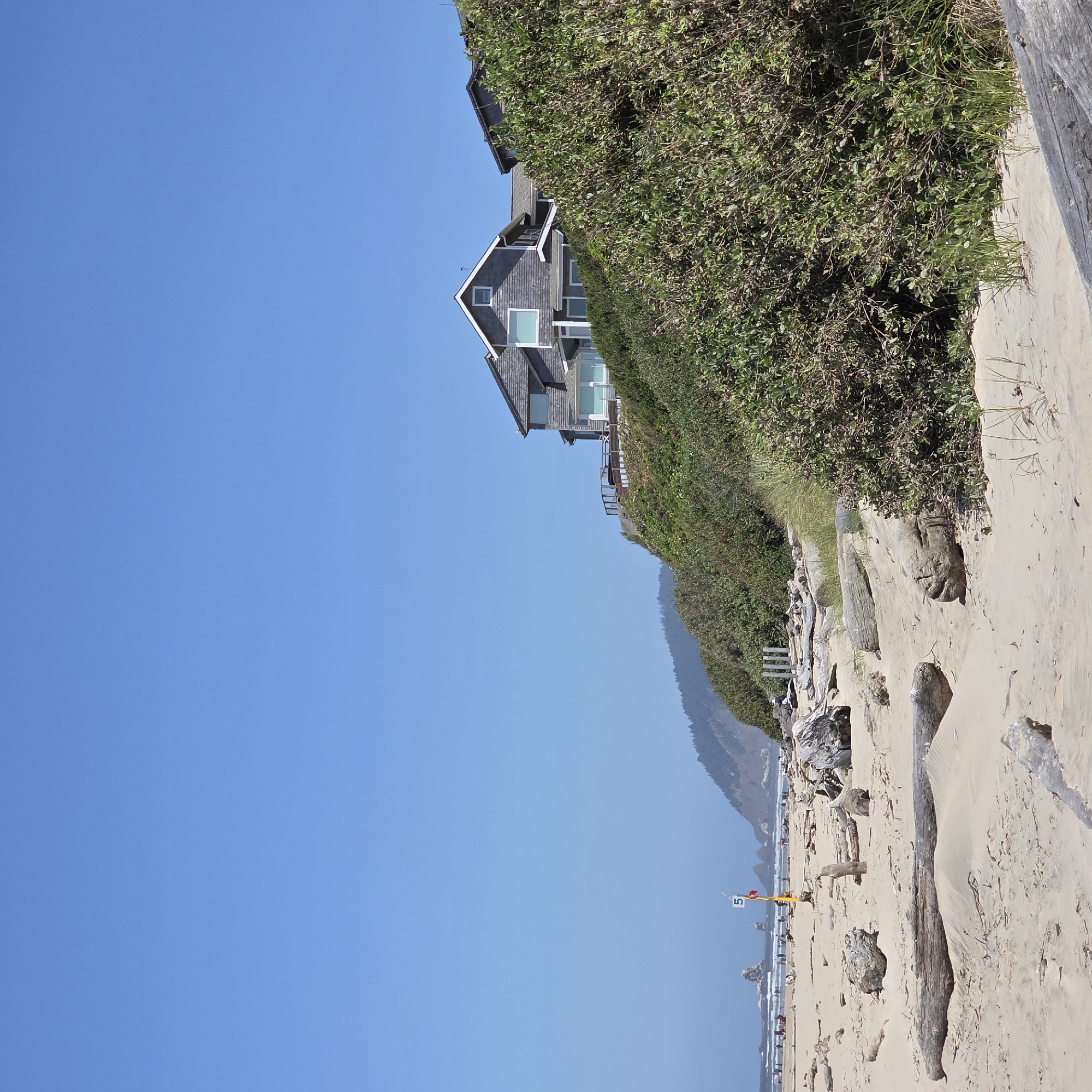 The image shows a beach scene in Cannon Beach, Oregon, United States of America. In the foreground, there is a sandy beach with various pieces of driftwood scattered across it. The sand appears to be somewhat damp and rippled in places. Further back, a lush green hillside is covered with dense vegetation. Atop the hillside, a large, multi-story house with a gray shingled exterior and multiple windows is visible. The house has a deck with railings, and parts of its roofline and chimneys are also discernible. The sky above is a clear, bright blue, with no clouds present. In the distance, the ocean can be seen with waves and rocky outcrops. A few people are visible on the beach and in the water in the far distance. A yellow pole with a red and white flag and a sign displaying the number "5" is also present on the beach. The overall impression is a sunny day at the coast with a prominent house overlooking the beach.