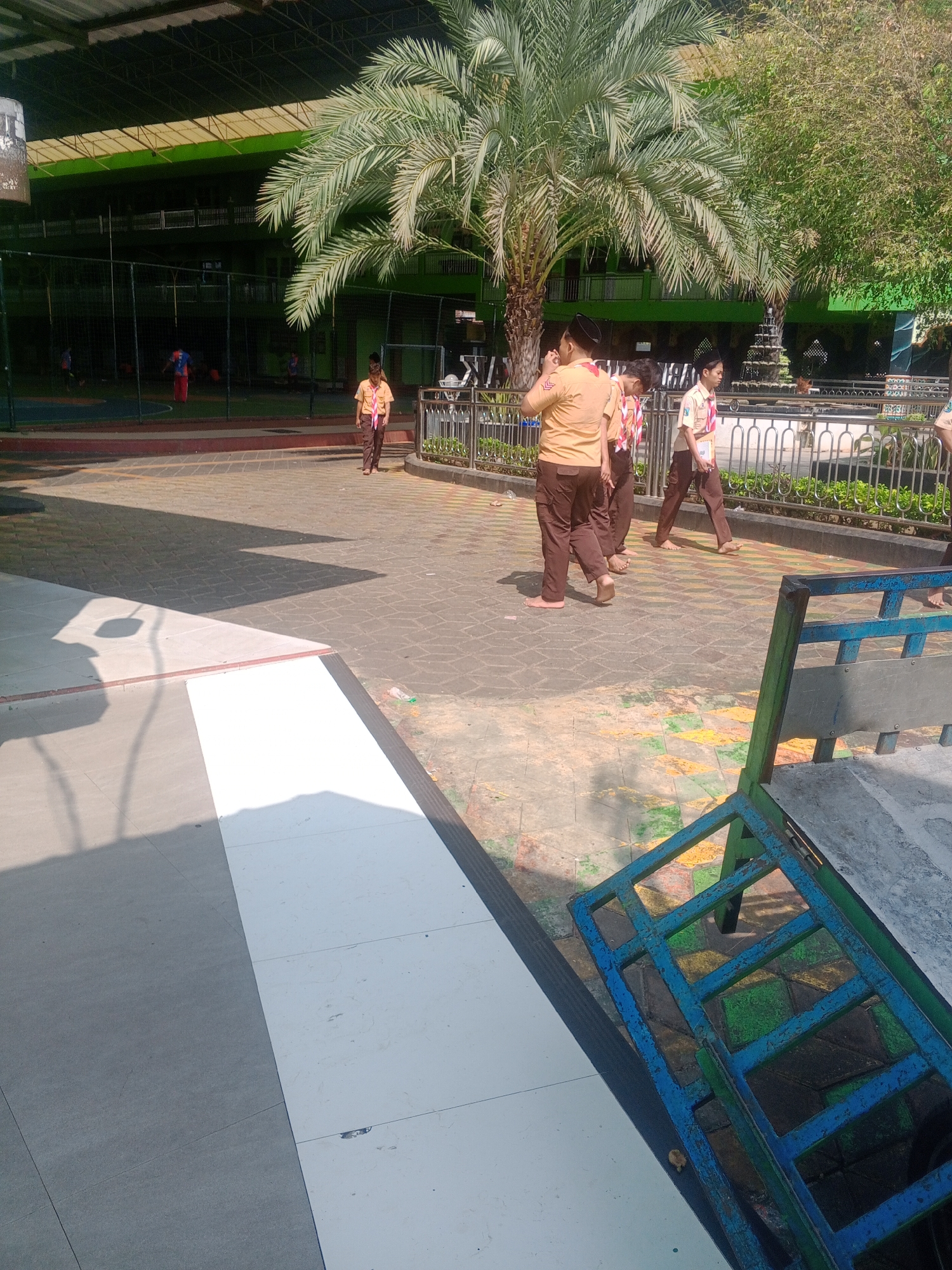 In Ponorogo, Indonesia, on a paved outdoor area with a patterned tile surface, a group of young people in scout uniforms are walking. The individuals are wearing tan shirts and brown pants, and at least two are barefoot. A large palm tree is visible in the background, as is a green building with a dark roof structure supported by a visible framework. The scene appears to be a school or public facility, with a fence and greenery bordering a walkway. A blue metal structure, possibly a bench or part of playground equipment, is in the foreground, casting a shadow on the ground. Sunlight casts distinct shadows, indicating a clear day.