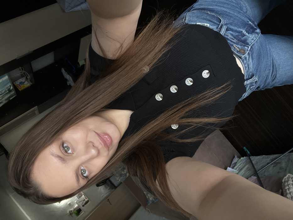 A female individual is depicted in a close-up selfie taken within an indoor residential setting in Chaikovsky, Russia. She possesses long, straight, brown hair, green eyes, and pink lips, which are slightly parted in a faint smile. She is wearing a black ribbed short-sleeved polo top with a collar and five visible white circular buttons down the front, paired with blue denim jeans. Her left arm is partially visible on the lower left of the frame, indicating the selfie action. The background on the left features a light-colored wall with mounted shelves displaying small plants and decorative objects, alongside a partial door frame and a light-colored textile item on the floor. The background on the right is darker, showing a rectangular wall-mounted artwork with blue tones, a shelf holding a Lipton tea box and other items, and scattered clothing on a surface beneath it. The floor visible is dark-colored, possibly wood.