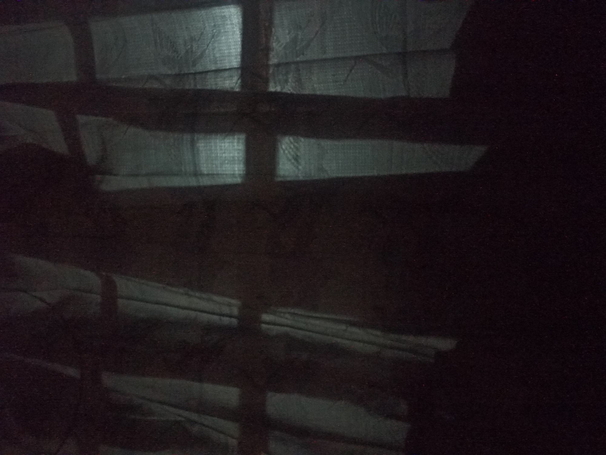The image shows a dark, blurry view through a window with curtains. The window panes are visible as vertical and horizontal lines casting shadows on the fabric. The overall scene is dimly lit, suggesting it might be nighttime or a dark interior. There is no indication of the location within Tuba, Indonesia. The image lacks any clear details about people, subjects, objects, or actions. The environment appears to be indoors, with a window and curtains being the only discernible elements.