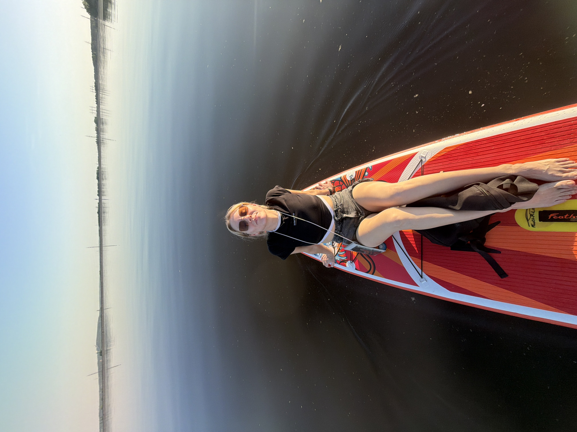 A woman wearing sunglasses, a black top, and gray shorts is lying on her back on a paddleboard, which is floating on water. The paddleboard is red and orange with white accents, and a black bag is placed on it. The water is dark, reflecting the sky. In the distance, along the horizon, are land and structures resembling power lines or pylons. The image is potentially taken in or near Noviy, Russia, due to the presence of structures reminiscent of those found in the area. The woman appears to be relaxing on the water.
