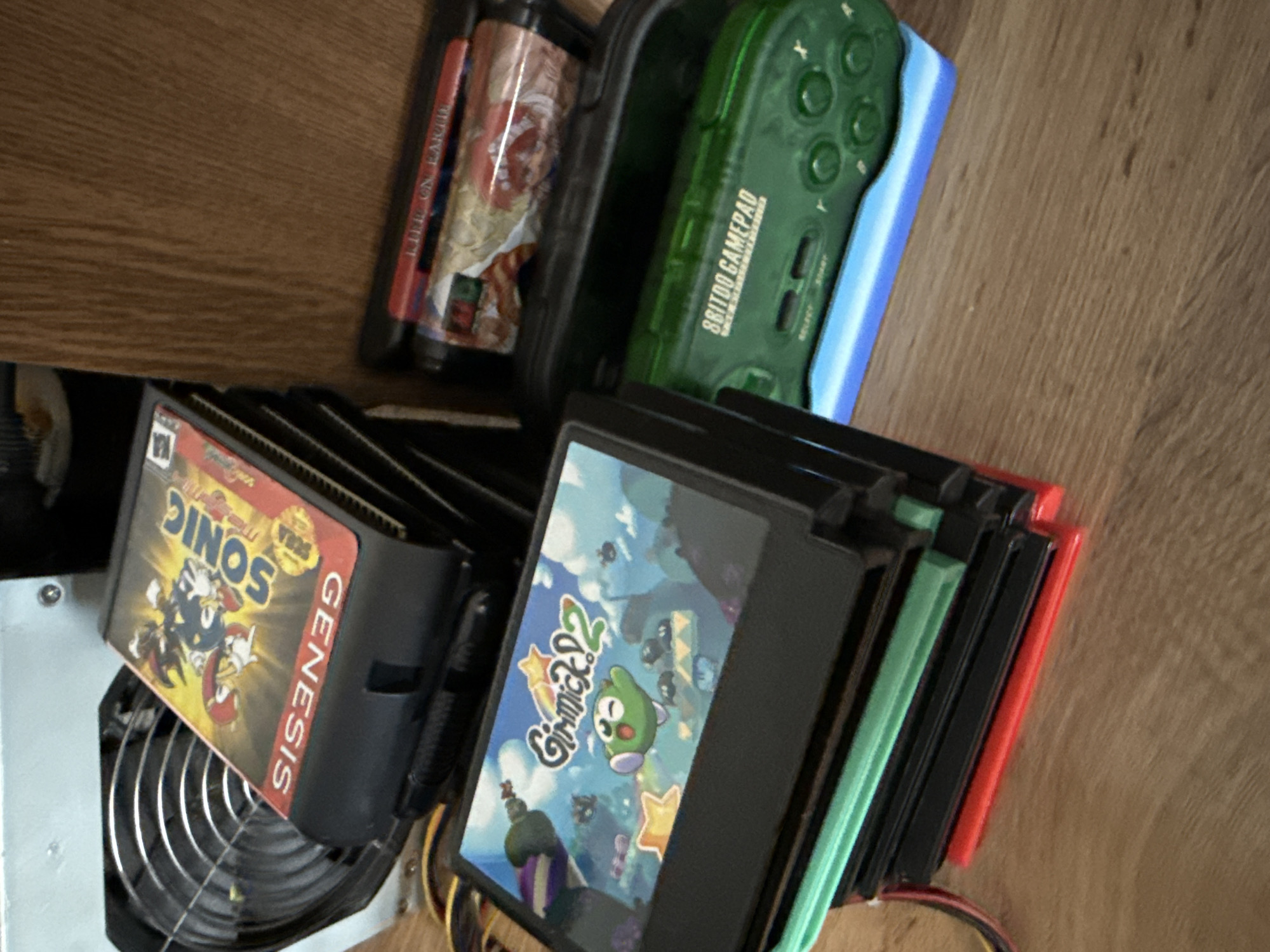 The image shows a variety of video game cartridges and accessories scattered on a wooden surface, likely a table or floor.
Objects:
- Several Sega Genesis game cartridges, including "Sonic 2", and "Gimmick 2" and other titles with visible red, green, and black labels. The game cartridges are stacked.
- A black cassette case holding something resembling rolls of film.
- A transparent green 8Bitdo gamepad.
- Metal fan.
- Wires
Environment:
- Wooden surface (table or floor).
Location:
- Unknown, not discernable from the image. The presence of video games does not indicate the location is Moscow, Russia.