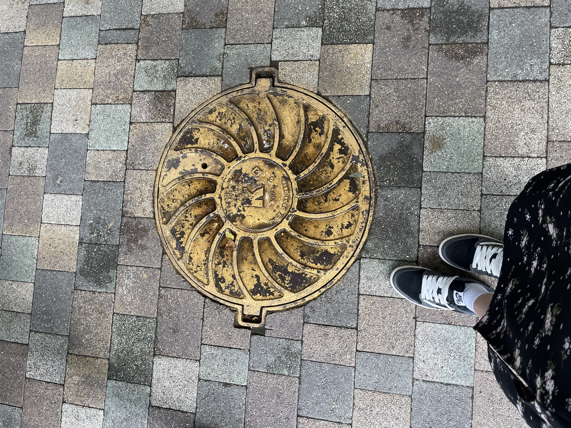 A manhole cover is centrally located on a paved sidewalk. The manhole cover is circular and made of metal, painted a distressed yellow color with a spiraling pattern and markings. The sidewalk is composed of rectangular pavers in various shades of grey and beige. A person's feet, wearing black and white sneakers and a dark floral skirt, are visible in the lower left corner of the image, standing on the sidewalk. The scene appears to be outdoors, likely in an urban environment. The location is not Stavropol, Russia.