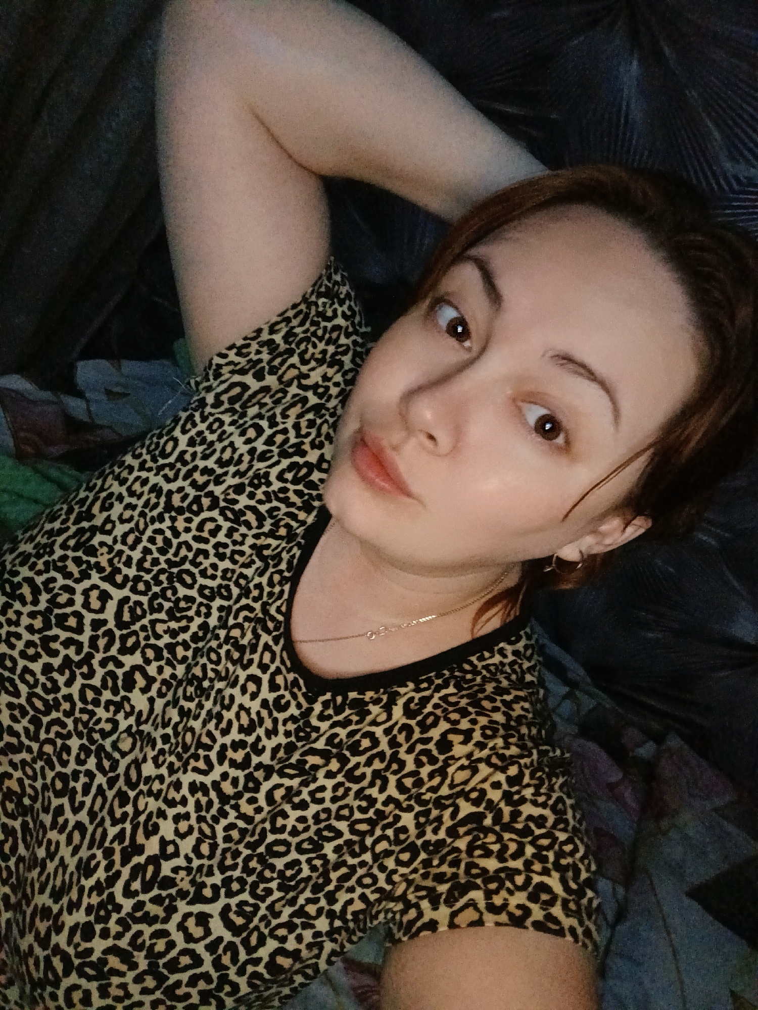 A woman with reddish-brown hair is lying on a bed. She is wearing a leopard-print shirt and a gold necklace. Her right arm is raised above her head. The bed has a dark, possibly leather, headboard and a patterned bedspread. The location is Chaikovsky, Russia.