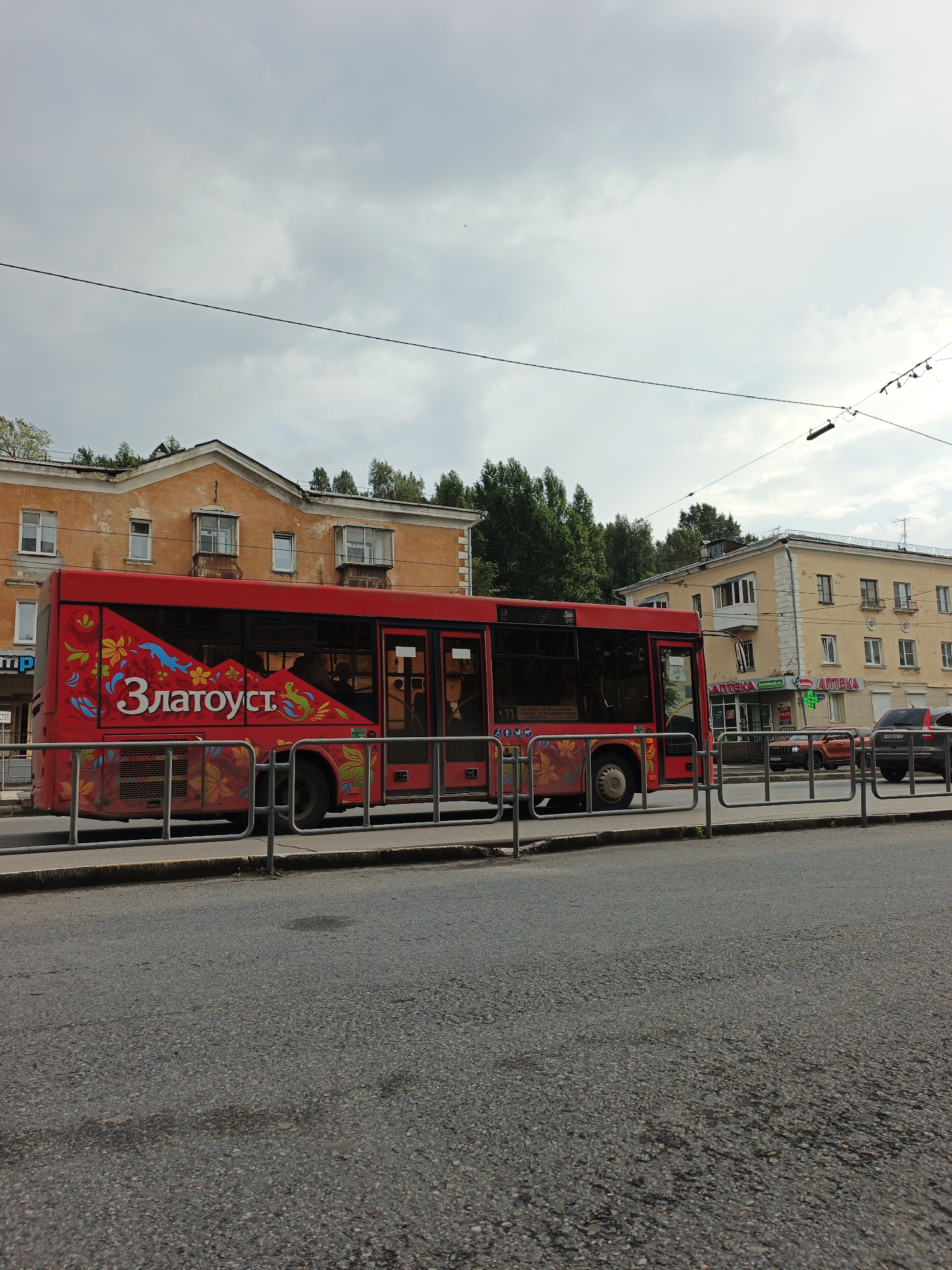 A bright red bus with floral and animal decorations is parked on a street in Zlatoust, Russia. The bus has "Златоуст" (Zlatoust) written in large white letters on its side, along with images of flowers, dolphins, and a lizard. The bus is on the right side of the image, with its side facing the viewer. In the background, there are two older buildings with yellow and orange facades, and trees. A metal fence runs along the sidewalk in front of the bus. The sky is overcast. Several cars are visible parked in front of the yellow building on the right, including a dark-colored SUV. There is a pharmacy with green signage on the ground floor of the yellow building.