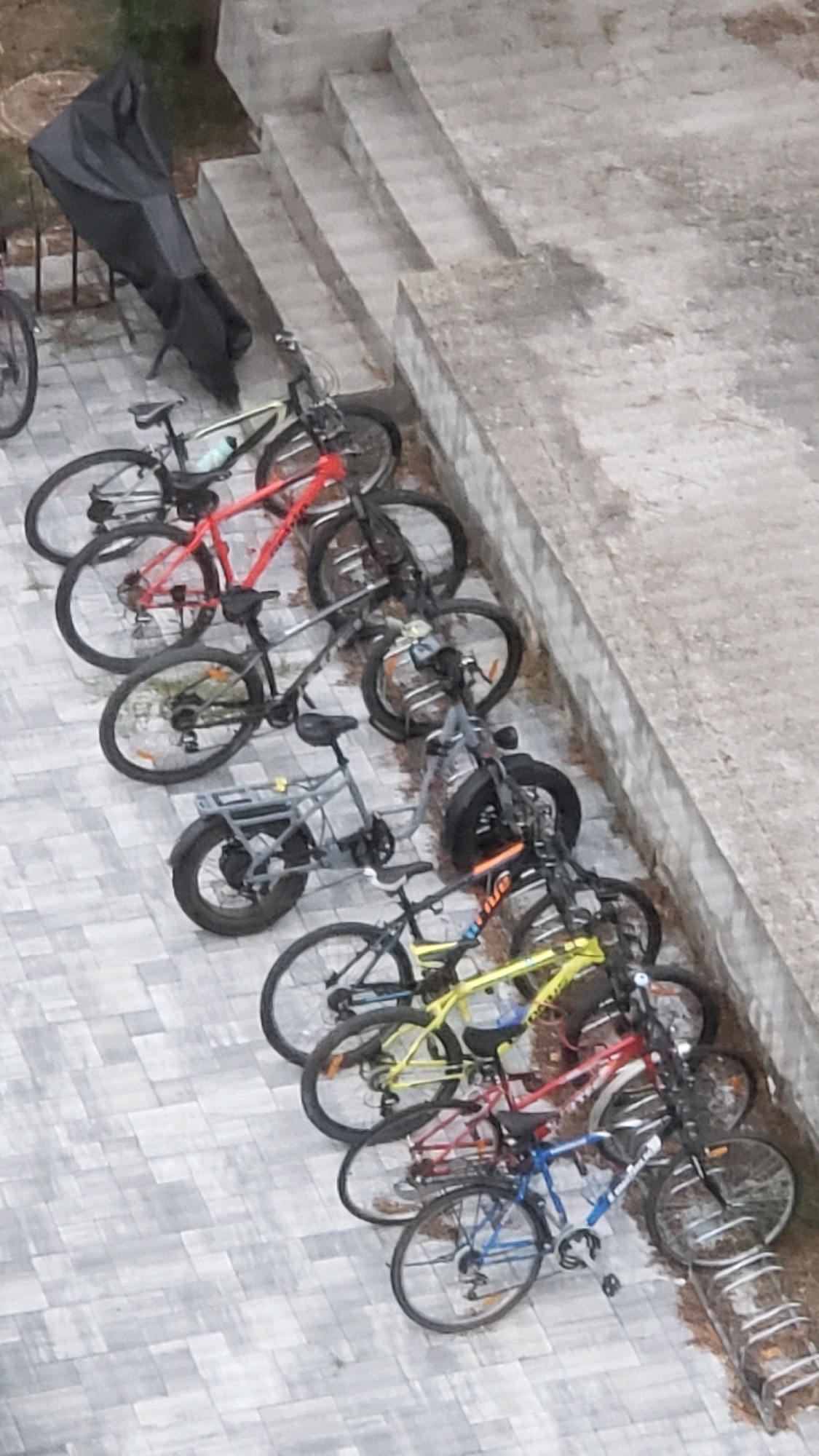 A high-angle, medium shot captures a row of ten bicycles parked on a patterned grey tiled ground, alongside a concrete structure with steps. The bicycles are lined up, some leaning against each other, suggesting a communal parking area. From left to right, the bikes appear to be: a black bike partially covered by a black tarp, a dark blue and grey bike, a red bike, a grey bike, a grey electric bike with a prominent rear rack, a black and orange bike, a yellow bike, another red bike, a blue and silver bike, and a black bike with silver accents. The environment appears to be outdoors, possibly a residential area or public space in Prešov, Slovakia, indicated by the tiled ground and concrete steps. No people are visible in the frame. The lighting suggests an overcast day.