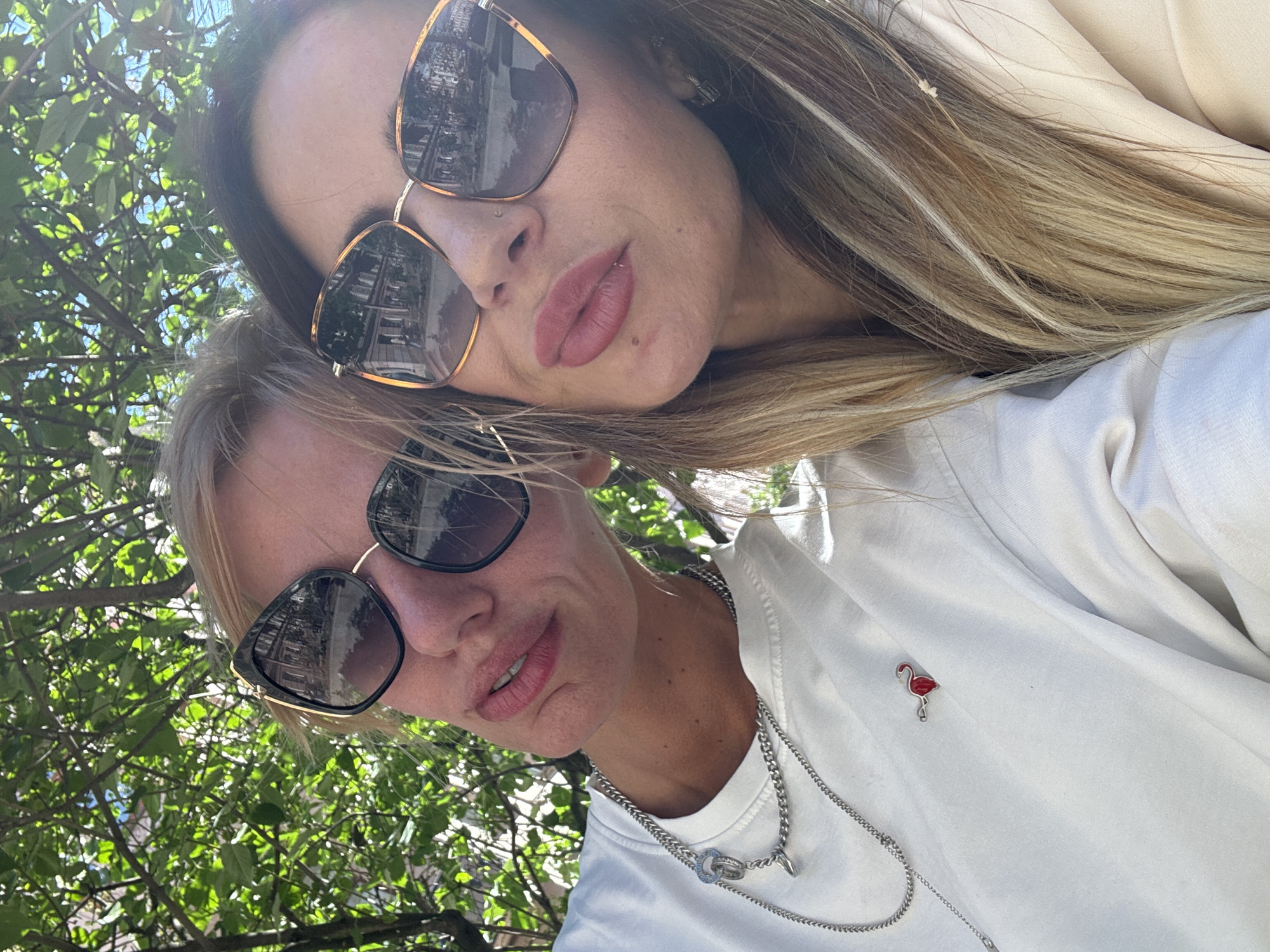 In a close-up selfie taken outdoors, two individuals are pictured with their faces angled towards the camera. The person in the foreground, on the right, has blonde hair and wears large, dark-rimmed sunglasses. They are sporting a white t-shirt adorned with a small, red flamingo pin and a silver chain necklace. Their lips are slightly parted, and they are smiling. The second person, in the background, has darker hair with blonde highlights and also wears oversized sunglasses with a gold frame. Their face is tilted, with their lips appearing to be pursed.
The background is filled with lush green foliage, suggesting an outdoor setting, possibly a park or garden. The lighting indicates it is daytime, with sunlight filtering through the leaves. The reflection in the sunglasses of the person in the foreground shows a street with buildings, hinting at an urban environment. The overall mood appears casual and happy, typical of a selfie taken with a friend. No visible text is present in the image.