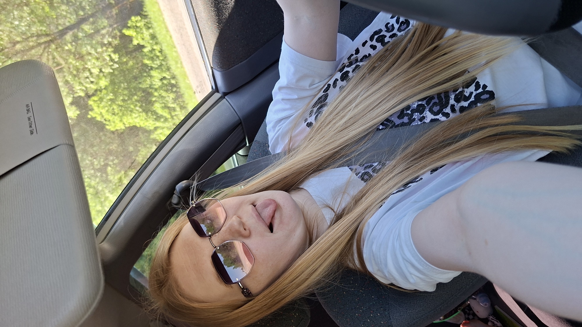 A young woman is in the passenger seat of a car, wearing a white T-shirt with a leopard print, sunglasses, and a seatbelt. She is sticking her tongue out. The outside is a forest that is visible through the car window. The location is likely in the city of Chaikovsky, Russia.