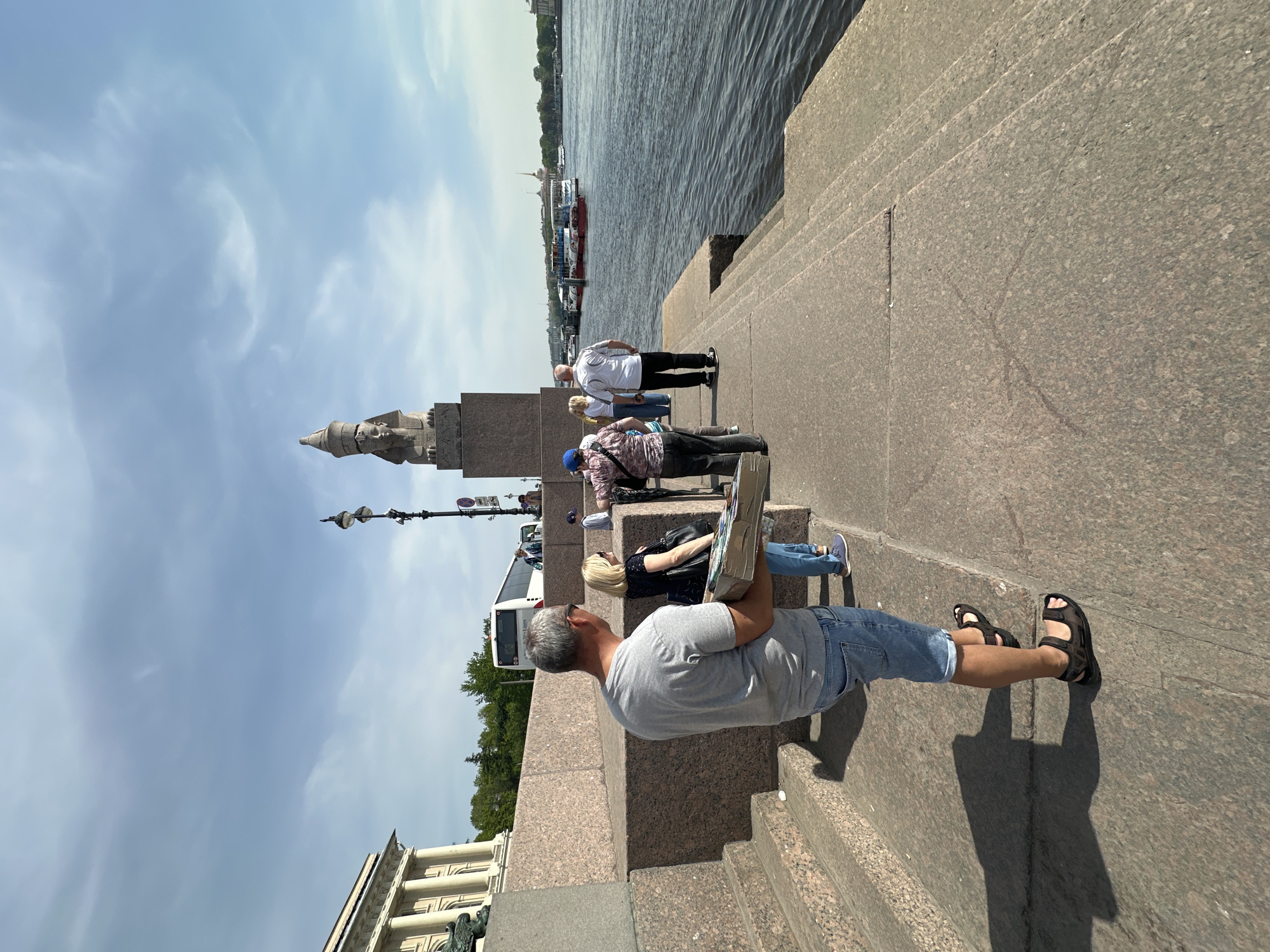 A wide-angle, outdoor medium shot depicts a scene on a granite embankment, likely in Saint Petersburg, Russia. In the foreground, a man is seated on the granite steps, facing away from the viewer, holding a painting board. He is wearing a gray t-shirt, blue shorts, and black sandals. Behind him, a group of people stands or sits on the steps. A white bus is parked nearby. A landmark, likely the Rostral Column of St. Petersburg, is visible against a blue sky with scattered clouds. In the background, the Neva River flows with several boats visible on the water. The scene is brightly lit, suggesting a sunny day. The perspective emphasizes the granite structures and the riverfront environment.