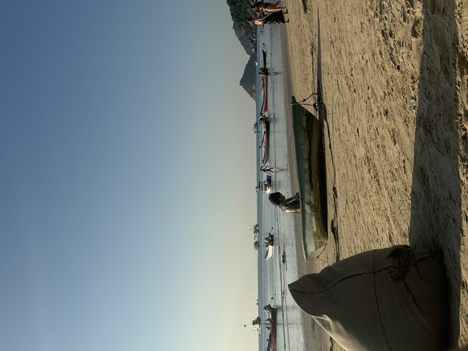A wide-angle photograph captures a bustling beach scene at the City of Unknown, Unknown, likely during late afternoon or early morning. In the immediate foreground, to the lower left, a large, light tan beanbag chair is partially visible, casting a long shadow towards the right. Centrally, a weathered green-grey boat is beached on the sand. An individual with dark, curly hair is seated within this boat, facing away from the viewer. To the right, two adults walk along the sandy shore; one wears a dark, sarong-like wrap, and the other is in light-colored swimwear. The calm ocean in the mid-ground and background is populated by numerous anchored and floating boats, including several longtail boats and larger vessels. Multiple individuals are visible in the shallow water, some walking. The horizon features a distant, lush green, hilly landmass on the right. The sky is clear and light blue.