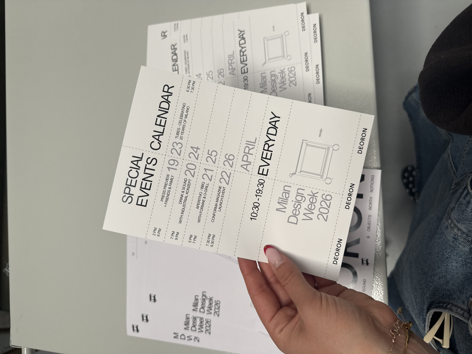 A person holds two tear-off promotional calendars with black ink. The calendars list special events and the words “Milan Design Week 2026.” The calendar includes the word “DEORON.” The woman is wearing jeans, a black shirt or sweater, and a gold bracelet.