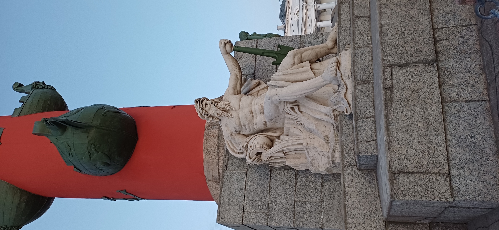 A low-angle, exterior, eye-level medium shot captures a monumental stone sculpture depicting a bearded male figure sitting on a tiered, grey granite base against a clear, pale blue sky. The sculpture is positioned in front of a thick, bright red cylindrical column that is adorned with green, ship-like decorative elements. The seated figure is draped in a white cloth that covers his lower body and left arm. His right arm is extended, holding what appears to be a trident or spear. He holds a cylindrical object in his left hand, from which water seems to be pouring down. The sculpture's surface shows some signs of age and cracking. Behind the main sculpture, a portion of another, smaller green sculpture is visible on the right. Further in the background, a white classical building with columns and windows is partially visible. The overall scene suggests an outdoor monument or architectural feature in a public space. This monument is the Rostral Column in St. Petersburg, Russia.