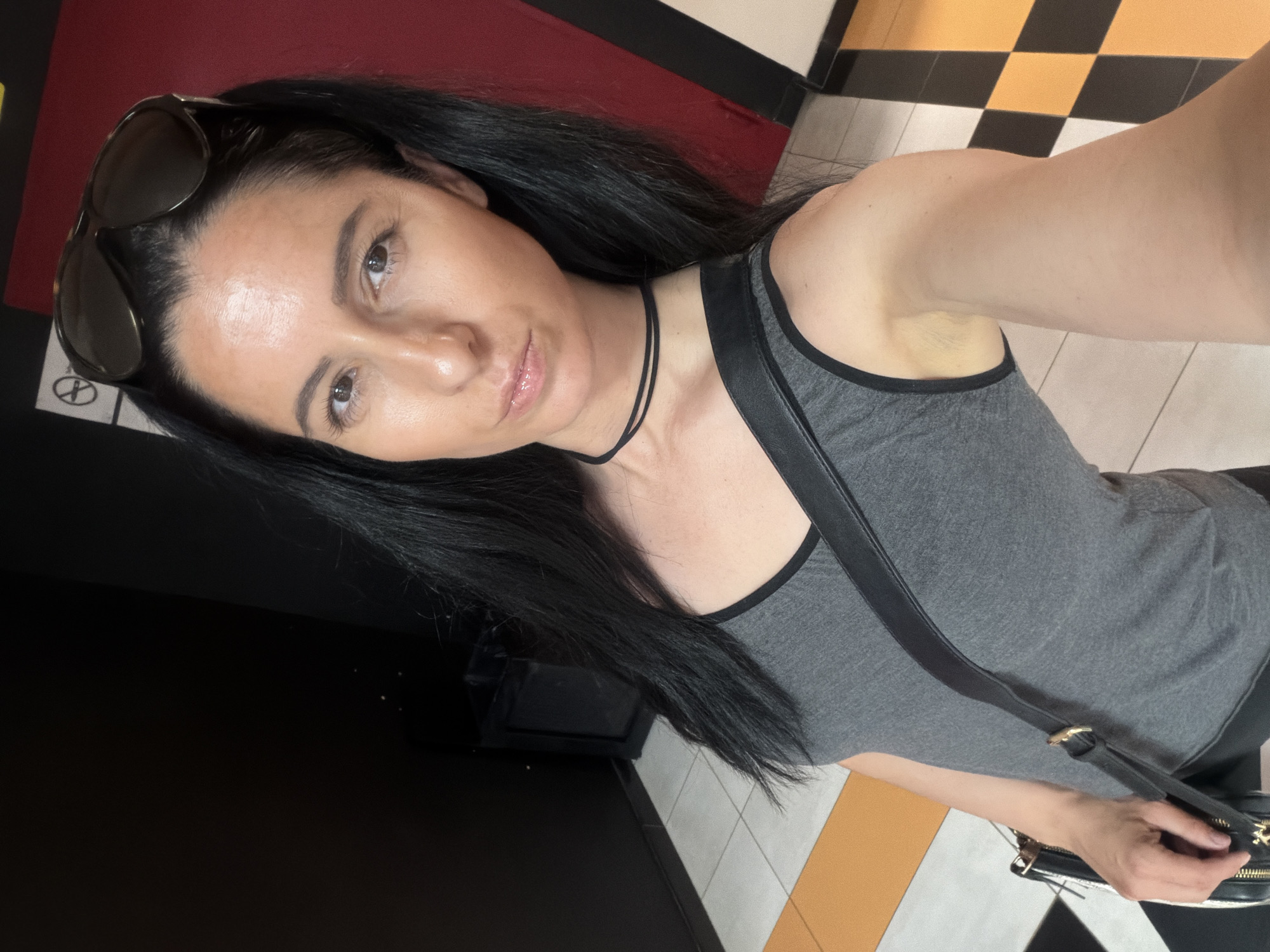 A woman with long, dark hair and sunglasses perched on her head takes a selfie. She is wearing a grey tank top with a black trim and a black choker necklace. A black purse with a gold buckle is slung across her body. The background appears to be an indoor public space with black, red, and black and orange checkered walls and floor. The location of Pleven, Bulgaria is not visually discernible in the image.