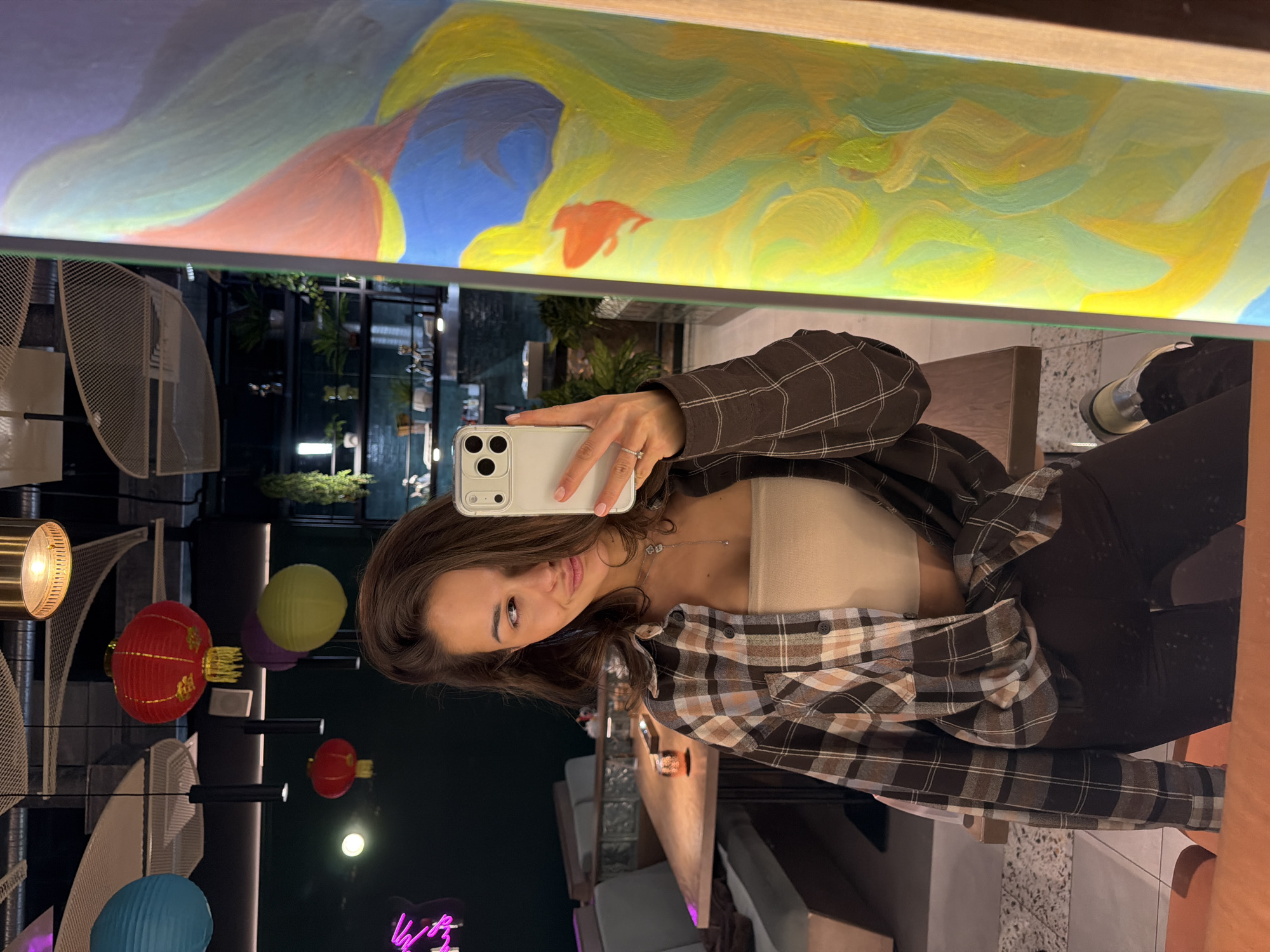 The image captures a young woman taking a mirror selfie in an indoor setting, likely an upscale cafe, restaurant, or lounge in Krasnoyarsk, Russia. She is positioned towards the right of the frame, holding a white smartphone in her left hand, which is reflected in the large mirror above her. She wears a brown and beige plaid shirt unbuttoned over a light beige tube top, with dark pants. A delicate necklace is visible, and a ring adorns her left ring finger. She looks directly at the phone's camera with a calm expression.
The upper portion of the image is dominated by the mirror's reflection of an abstract painting, featuring vibrant, fluid strokes in yellow, green, blue, and reddish-orange hues. Below the painting, the mirror reflects the woman and parts of the room's ceiling.
To the left and below the woman, the actual interior space is visible, characterized by modern design and ambient lighting. Several decorative paper lanterns in red, green, blue, and purple are suspended throughout the space, alongside various contemporary light fixtures, including mesh designs and a brass-colored sconce. Lush green plants are visible in the background, adding a natural touch to the contemporary decor. The lighting suggests the scene takes place in the evening or at night. A faint, glowing neon sign, mostly illegible but resembling "Vibes," is visible in the lower left corner. The floor has a light, speckled texture.