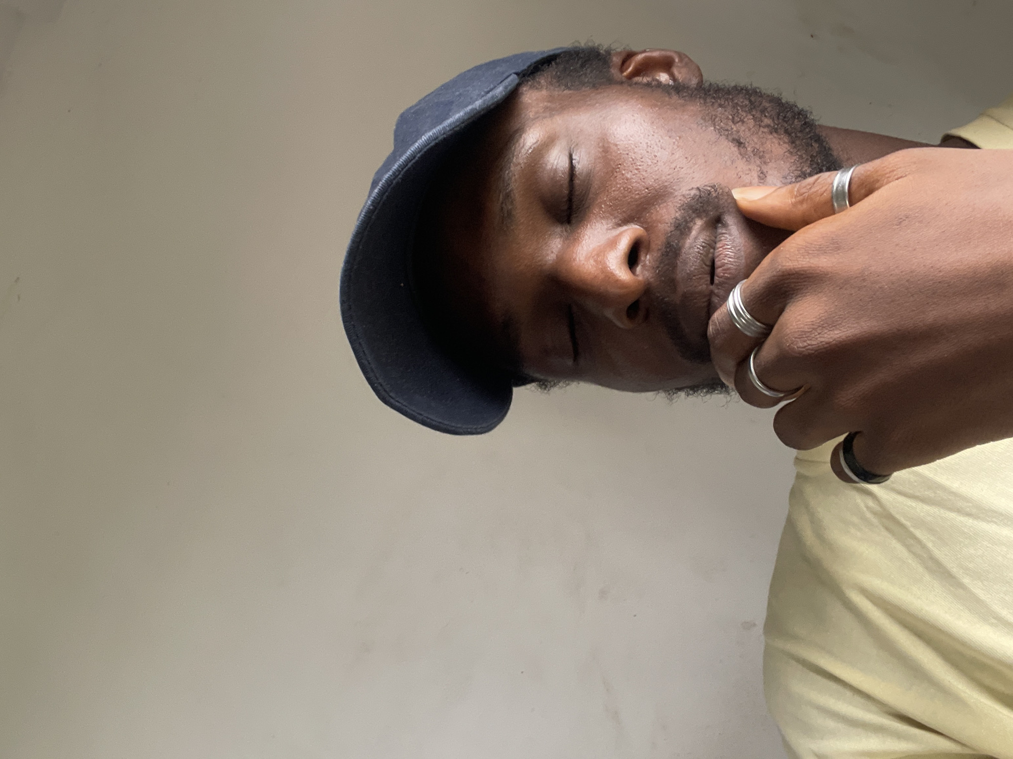 The image shows a close-up of a Black man wearing a navy blue baseball cap and a light yellow t-shirt. His eyes are closed, and his right hand is resting on his chin. He is wearing several silver rings on his fingers. The background is a plain, light-colored wall. The overall impression is one of contemplation or relaxation. The image was taken in Abuja, Nigeria.