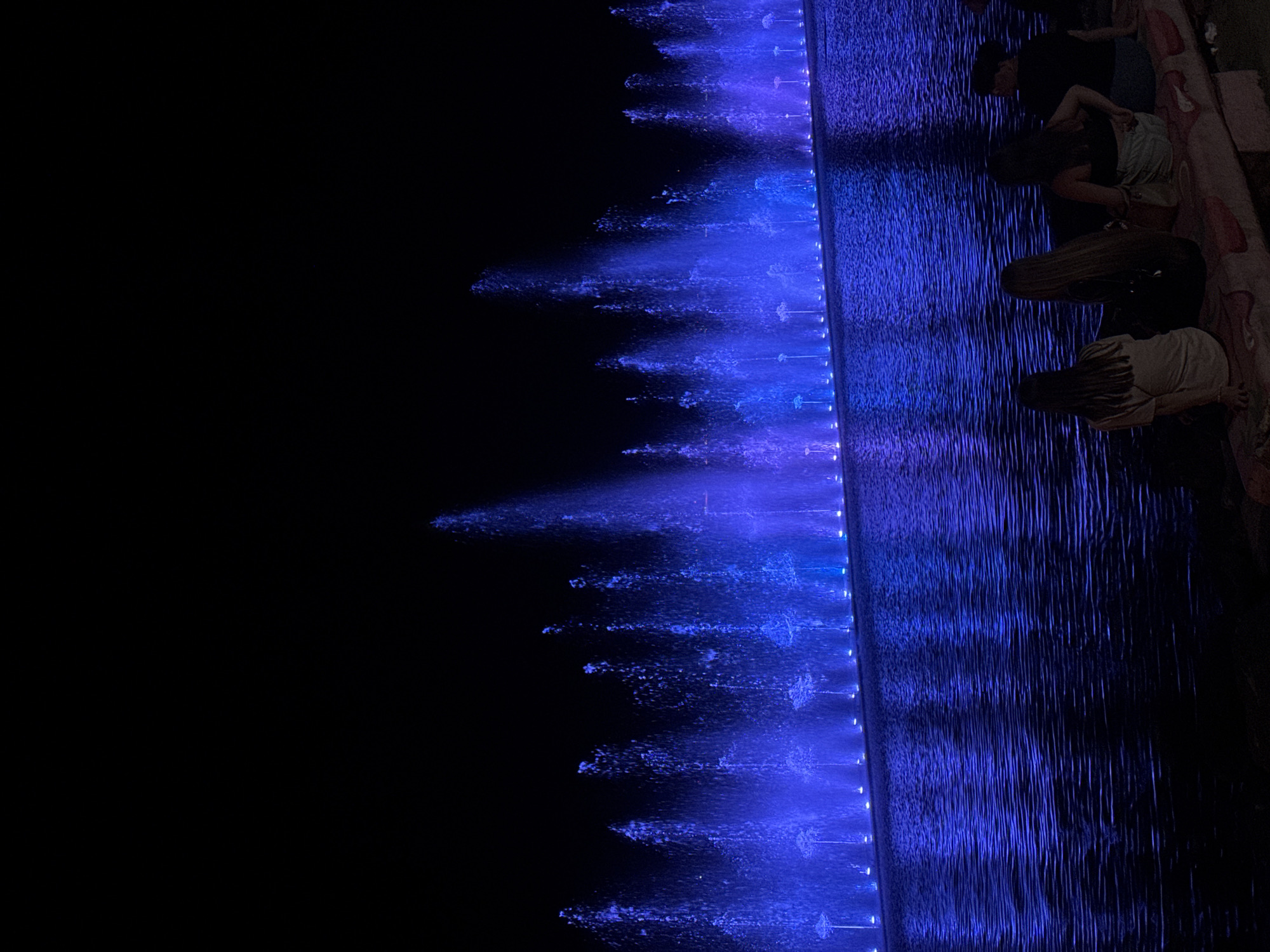 The image captures a nighttime view of a large water fountain display in Pomorie, Bulgaria, illuminated by vibrant blue lights. The fountain features numerous jets of water shooting upwards, creating a visually dynamic spectacle. The water surface reflects the blue light, creating a shimmering effect. In the foreground, four individuals are seated on a bench, facing the fountain and appearing to observe the display. The background is dark, suggesting it is late at night. The scene is set outdoors, likely in a public park or promenade area in the city of Pomorie.