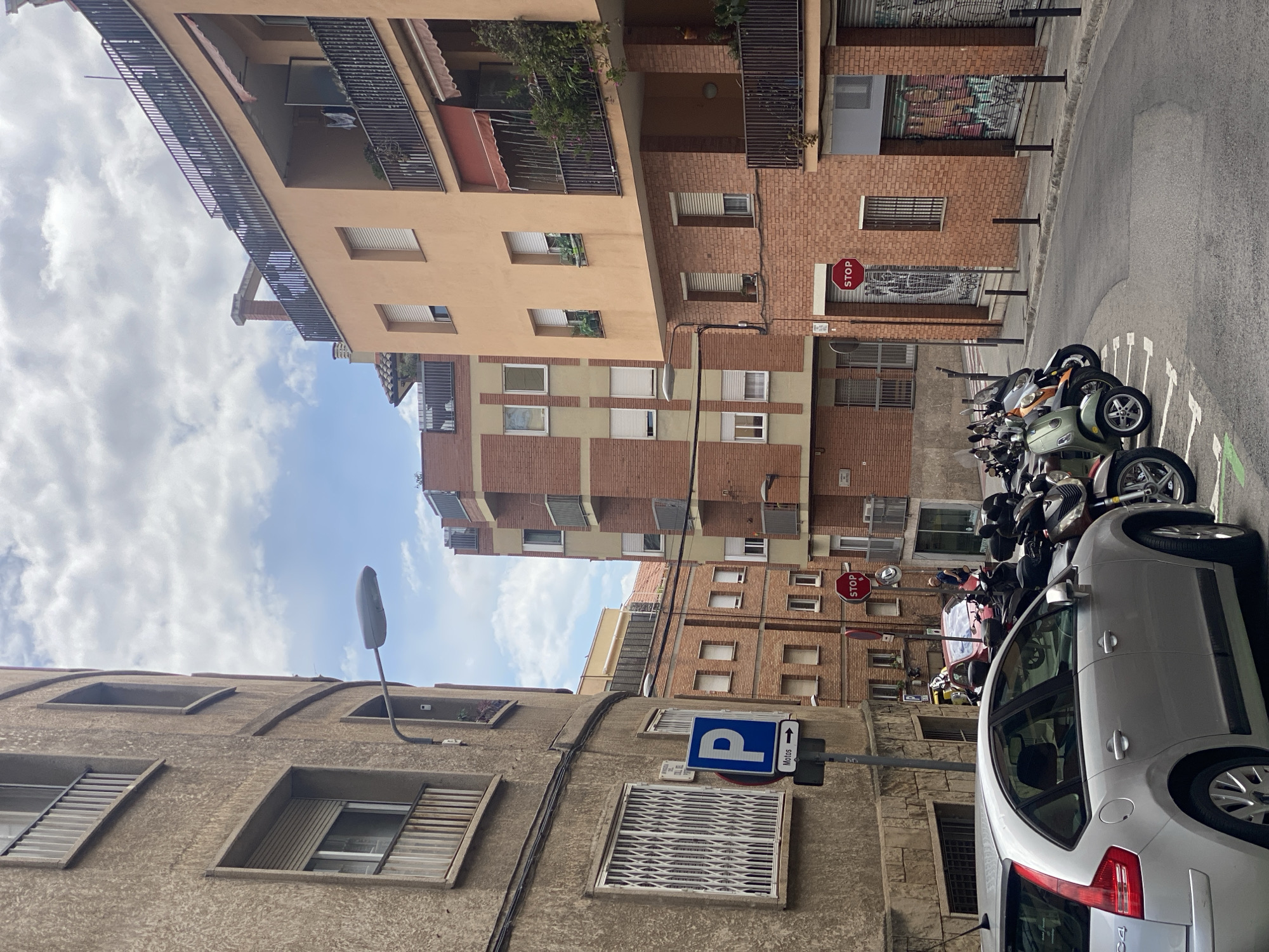 This daytime image depicts a typical urban street scene in Barcelona, Spain, under a partly cloudy sky. The street is flanked by several multi-story residential buildings with varied architectural styles.
On the left, a building with a rough, greyish-brown textured facade features multiple windows, some fitted with slatted blinds. A black street lamp post curves upwards from this side of the street. Prominently placed is a blue square "P" parking sign with a white arrow pointing up and the word "Motos" (motorcycles) printed below it.
The right side of the asphalt street is occupied by a line of parked motorcycles and scooters. In the foreground on the right, the rear and side of a silver hatchback car are partially visible. Beyond the parked vehicles, a red octagonal "STOP" sign stands out against a brick building facade. This building, along with others further down the street, showcases a mix of red brickwork and lighter painted sections. Some ground-floor windows or shop fronts on these buildings appear to be covered by metallic shutters, some of which are covered in colorful graffiti. Balconies with dark metal railings and some potted plants are visible on the upper floors of the buildings. The overall scene conveys a busy, lived-in city environment.