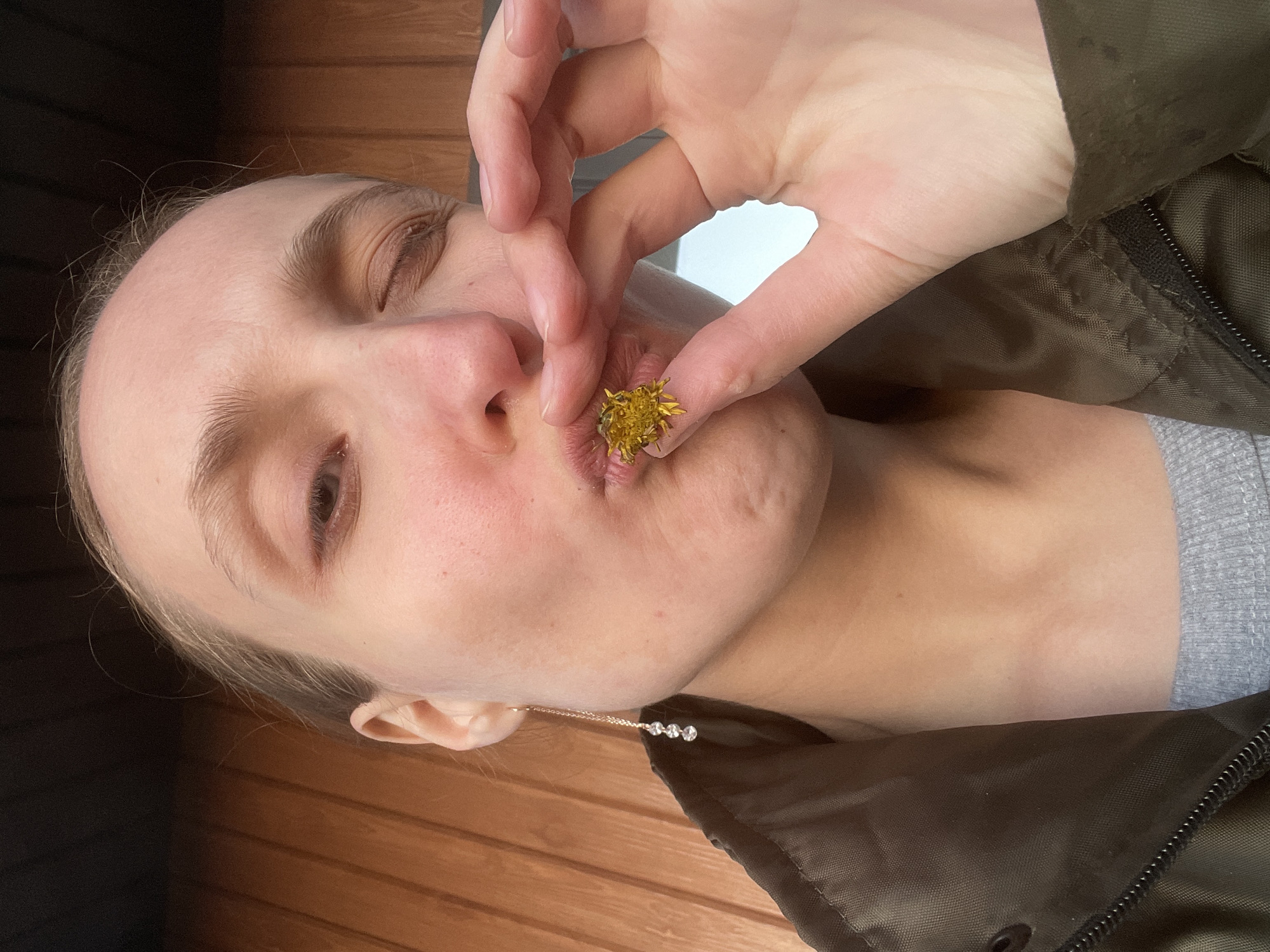 A woman is holding a yellow dandelion flower near her mouth. She is wearing a green jacket over a gray shirt. A delicate gold chain with four small, clear stones is visible near her ear. The background appears to be wooden planks. No information is available to determine if this picture was taken in the city of Подшивалово, Russia.