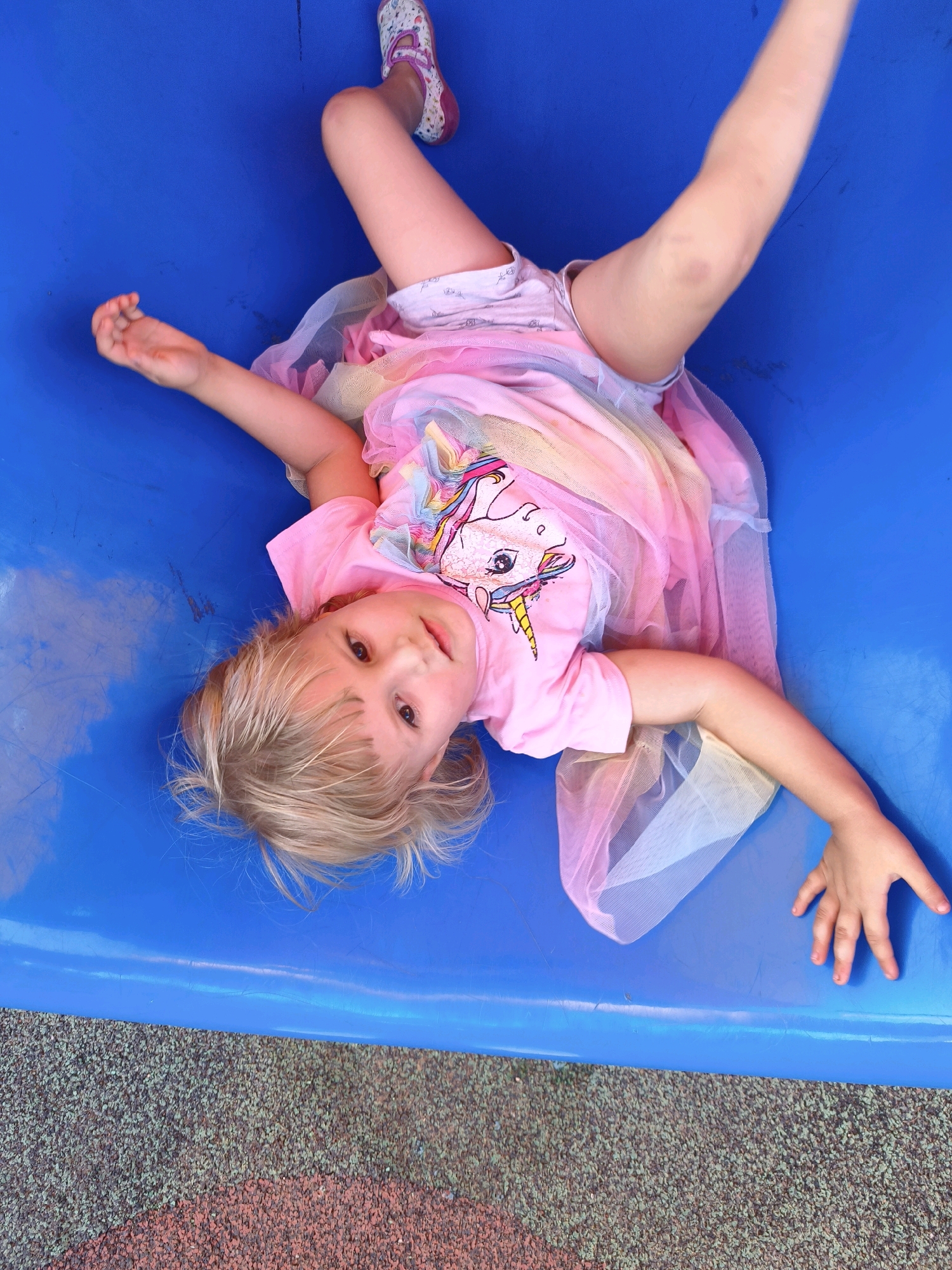 A young child with blonde hair lies on their back in a blue, plastic slide or play structure. The child is wearing a pink t-shirt with a sequined unicorn graphic, a multi-colored tulle skirt, and a white shoe with pink accents. One leg is bent, and the other is extended upwards, with the foot visible. The child's arms are spread out, with one hand reaching upwards and the other resting near their side. The child is looking up at the camera with a neutral expression. The background is the bright blue plastic of the play structure, with a textured, reddish-brown surface visible at the bottom, likely playground ground cover. The lighting suggests daytime, possibly outdoors. The image appears to be taken in the Lachish Regional Council, Israel.