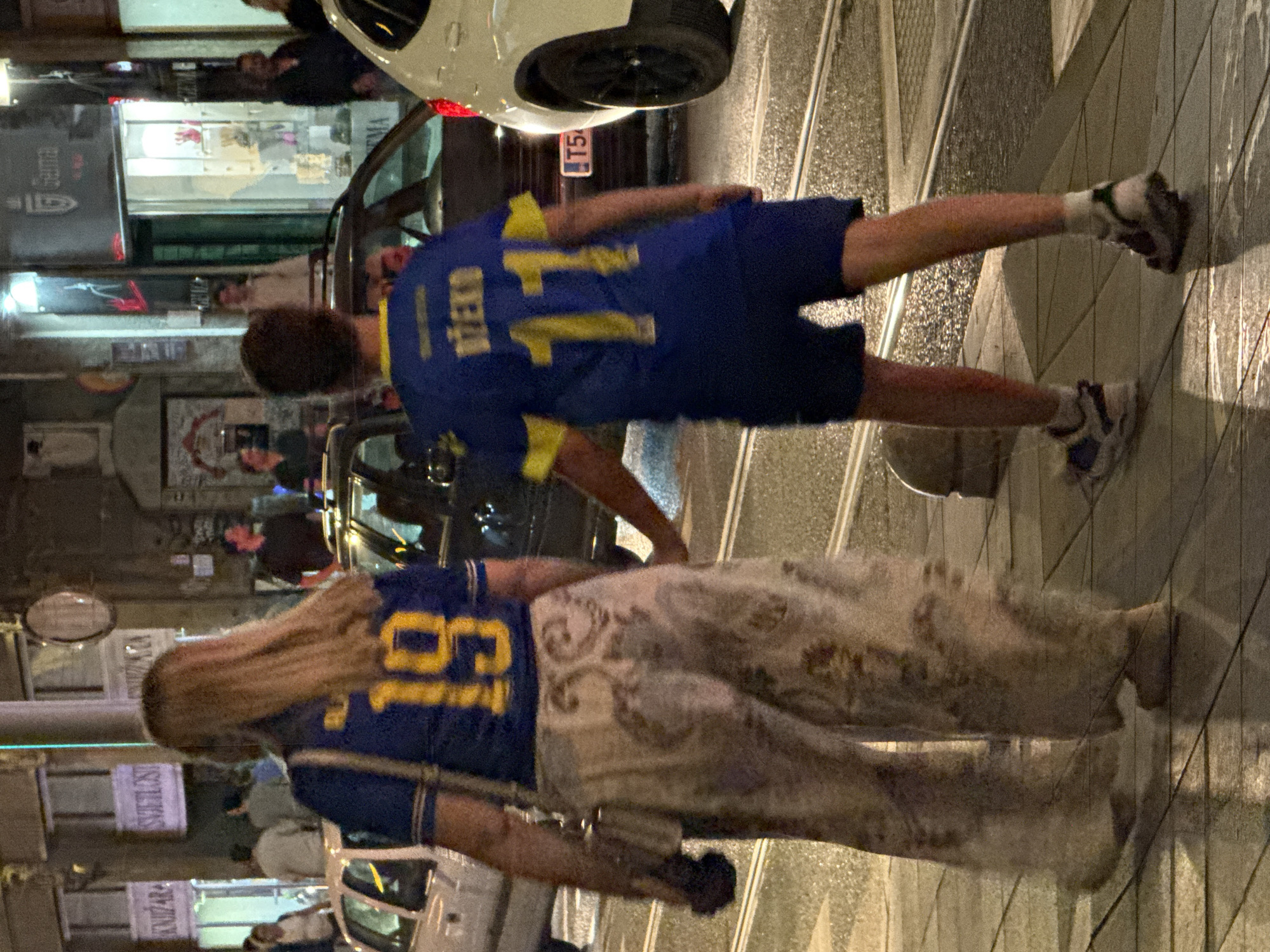 In Sarajevo, Bosnia and Herzegovina, at night, two individuals are captured walking on a wet city street. The individual on the left, a woman with long, blonde hair, is wearing a blue and yellow jersey with the number "19" prominently displayed on the back and a pair of loose, patterned pants. She is carrying a small, light-colored purse. The individual on the right, a youth, is wearing a blue jersey with "DŽEKO" and the number "11" on the back, paired with blue shorts and white socks with sneakers. They are walking on a street with tram tracks, and several cars are visible, including a white SUV partially in the frame on the right and a darker car to the left. The background shows storefronts and buildings with artificial lighting.