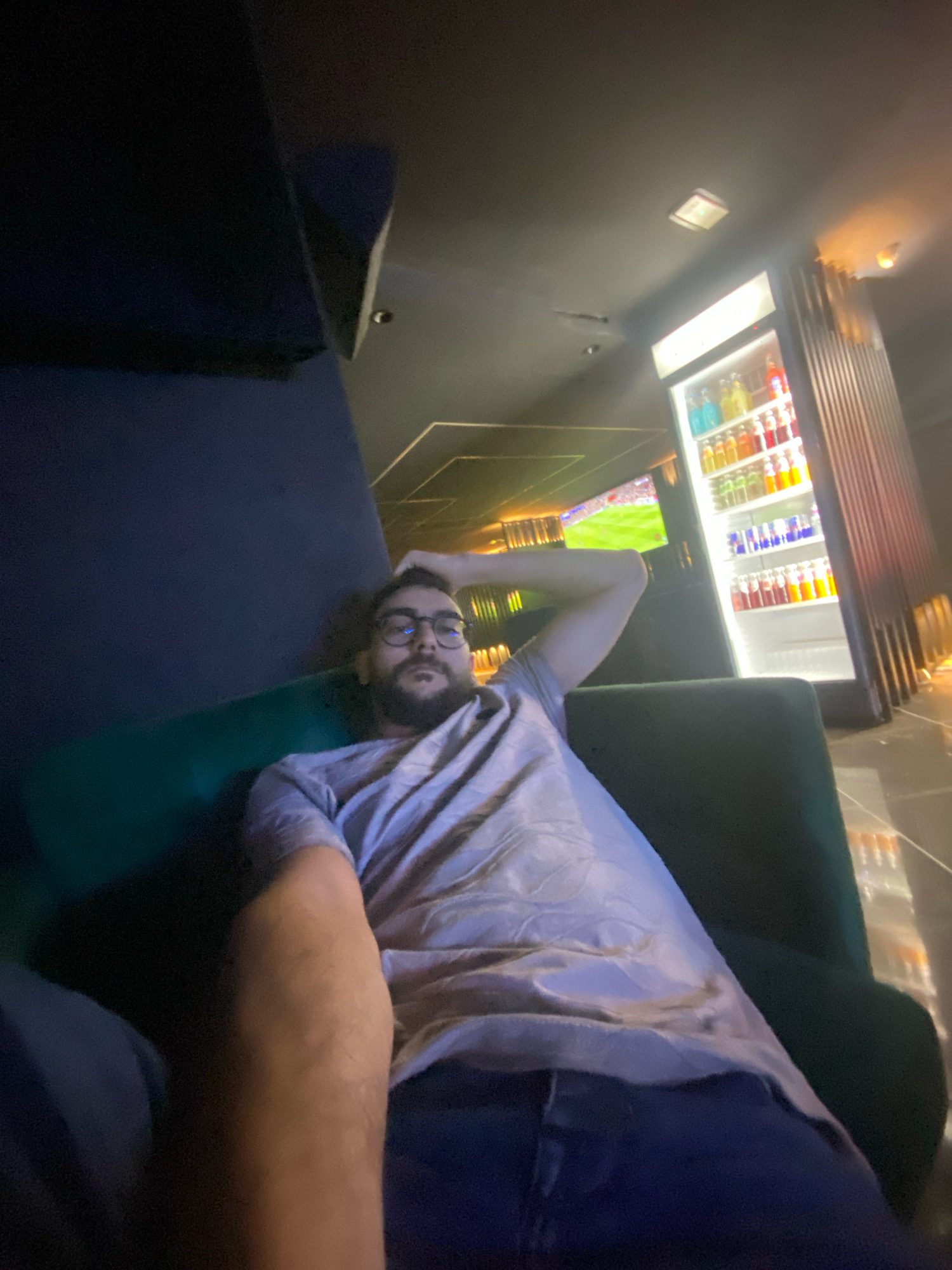 In Meknes, Morocco, a man with glasses and a beard is reclining on a dark green couch, resting his head on his arm. He is wearing a light purple t-shirt and blue jeans. Behind him, a brightly lit beverage cooler is stocked with numerous colorful bottles of drinks. To the left, a portion of a dark blue wall is visible. In the background, a television screen displays a sports game. The lighting in the scene is dim, with the cooler and television providing the primary light sources.