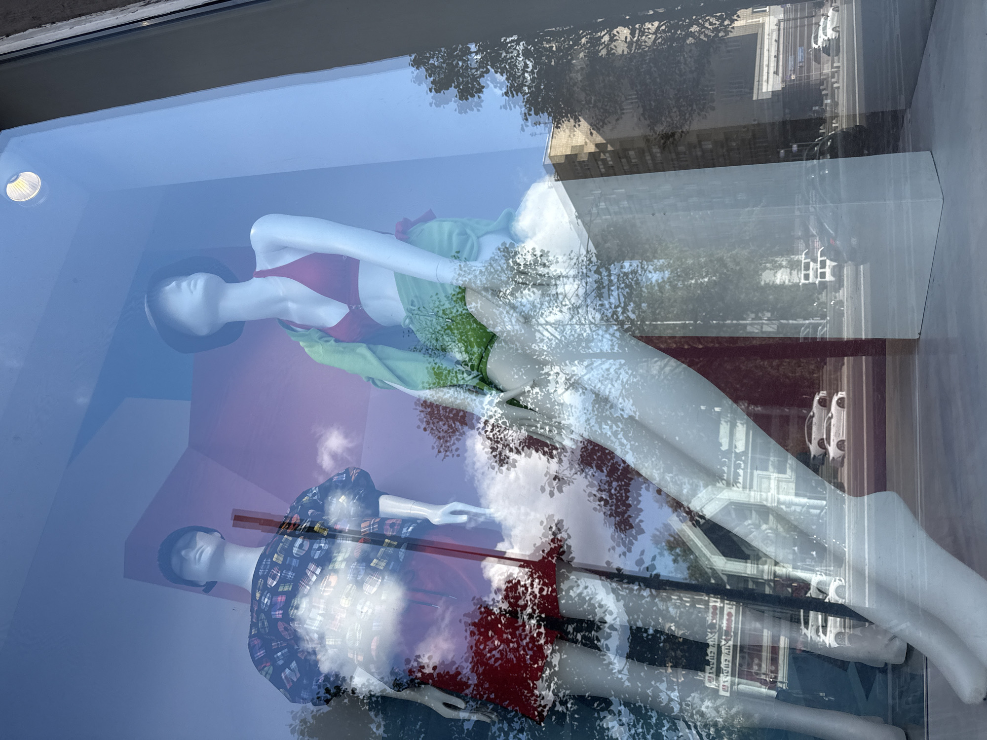 The image shows a reflection on a storefront window in Moscow, Russia. The reflection displays two mannequins dressed in clothing, possibly from the store. One mannequin is wearing a red bikini top and green shorts, and the other is wearing a plaid shirt and red skirt. In the background of the reflection, there are visible trees, buildings, and cars, indicating an urban environment. The sunlight is also reflected on the window, creating a bright and somewhat distorted view. The actual storefront interior is not clearly visible due to the reflections.