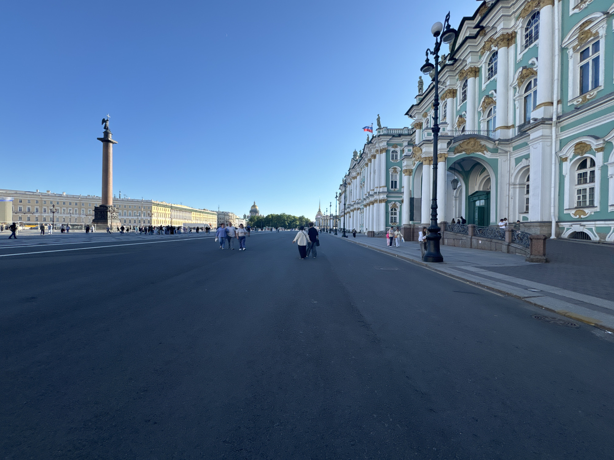 The image captures Palace Square in Saint Petersburg, Russia, under a clear blue sky. The scene is dominated by the expansive asphalt road in the foreground, leading towards a distant monument and buildings. On the right side of the image, the ornate, turquoise and white facade of the Winter Palace stretches along the square, featuring classical architecture with columns, arched windows, and decorative elements. A lamppost stands prominently in front of the palace.
On the left side of the square, the Alexander Column, a towering granite obelisk topped with a statue of an angel, is visible against the sky. Behind the column and to its left, a long, neoclassical building with a yellow facade forms the backdrop.
Several individuals are present on the square, walking and standing. Most are dressed in casual attire. They are scattered across the open space, with some groups walking towards the viewer and others walking away. The overall impression is one of a public space with visitors enjoying the architecture and open expanse. The lighting suggests it is daytime, likely morning or late afternoon due to the clear sky and lack of harsh shadows.