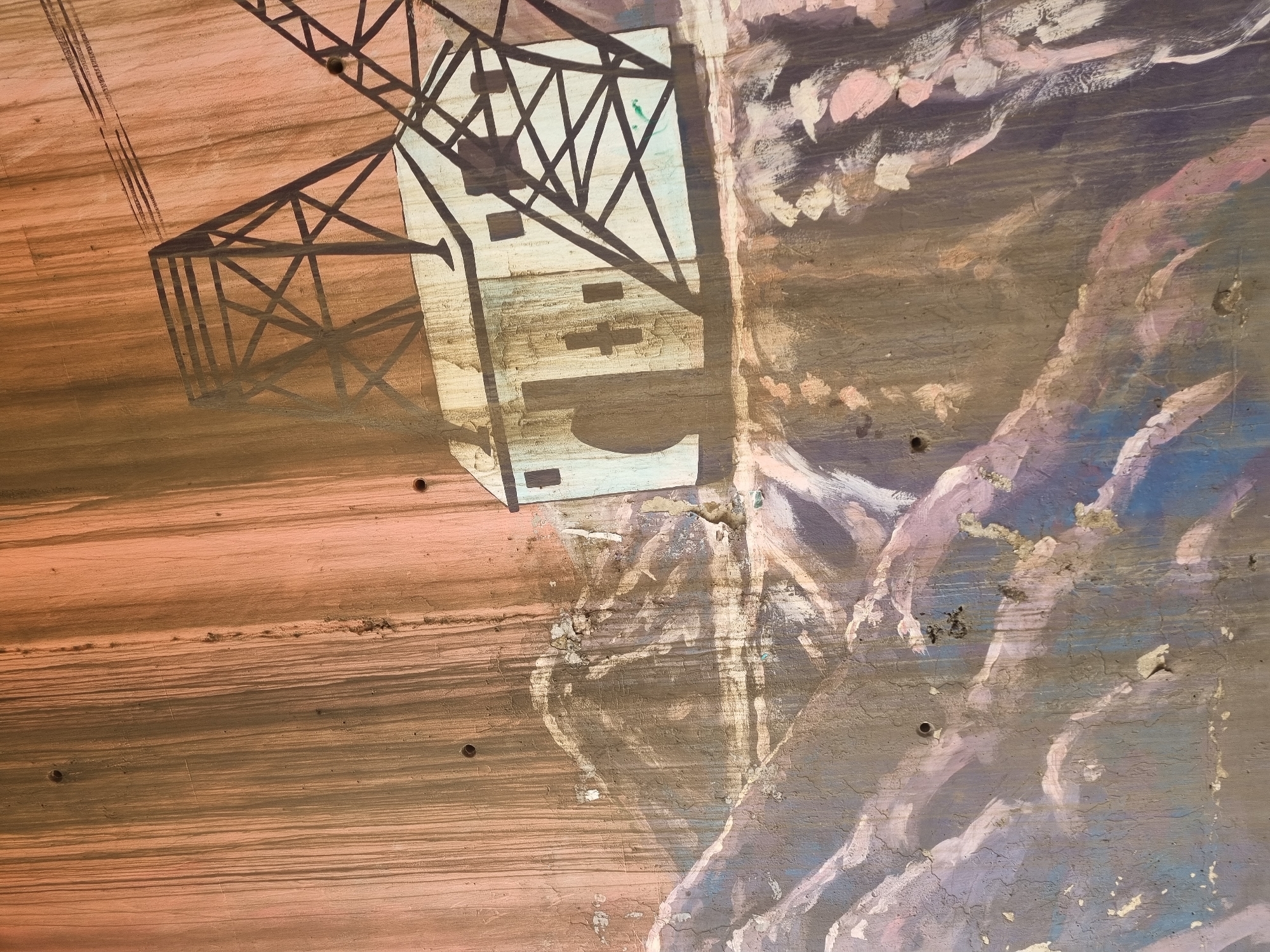 A close-up, partially obscured view of a mural on a weathered surface, possibly a wall. On the left, a detailed black rendering of a crane structure is visible against a backdrop of horizontal brown and peach hues, suggesting painted wood grain. The crane illustration casts a shadow effect over a lighter-colored, rectangular building-like form with dark details. To the right of the crane, the mural transitions to a different artistic style featuring swirling strokes of mauve, blue, and white, partially obscuring the crane. The surface exhibits signs of aging and wear, including cracks, peeling paint, and discoloration, along with several nail holes. There are no people visible. The location in Jos, Nigeria is not discernible from the image.