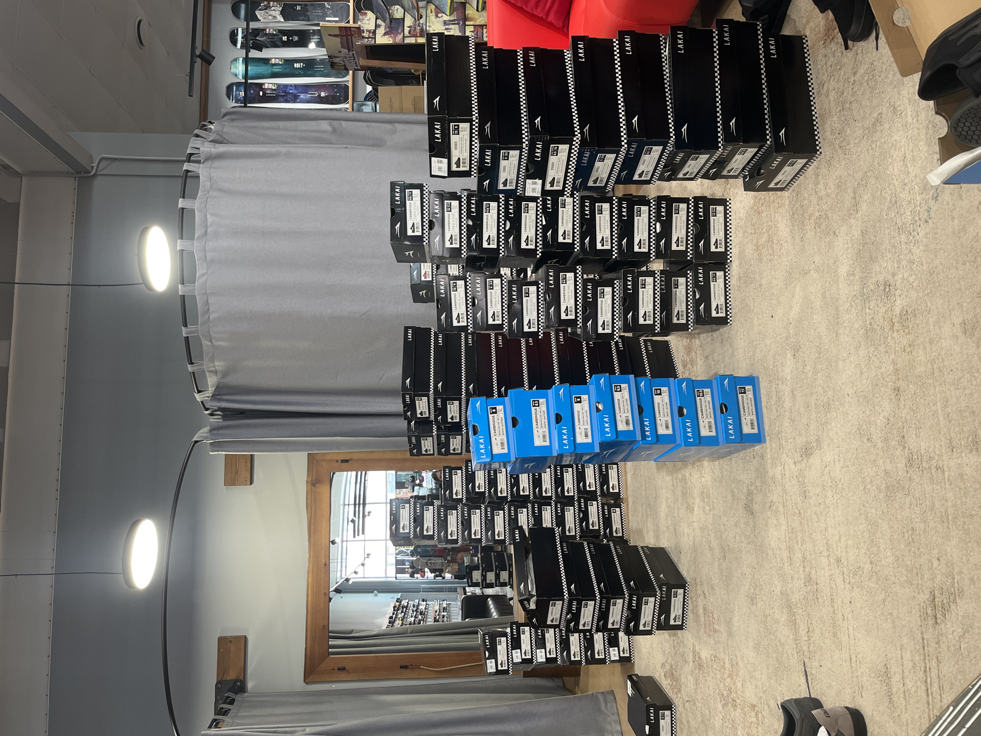 The image depicts a retail store interior, likely a shoe store or skate shop, with numerous stacked shoe boxes. The boxes are predominantly black with white branding that reads "LAKAI" and checkerboard patterns. There are also several stacks of bright blue boxes, also branded "LAKAI" and detailing shoe sizes and styles such as "CAMBRIDGE" and "ESSEX". The scene is cluttered with these boxes, some piled high on the floor, suggesting a recent delivery or a disorganized inventory. In the background, a fitting room area with grey curtains is visible, along with shelves stocked with snowboards. A red sofa is partially visible on the right side of the frame. The lighting suggests an indoor commercial space with overhead lights. There are no visible individuals actively interacting with the merchandise, though a person's head is just visible behind a stack of boxes in the center of the image. The floor is a light-colored, textured material, possibly concrete or a similar industrial flooring. The location is identified as a retail establishment in Krasnoyarsk, Russia.