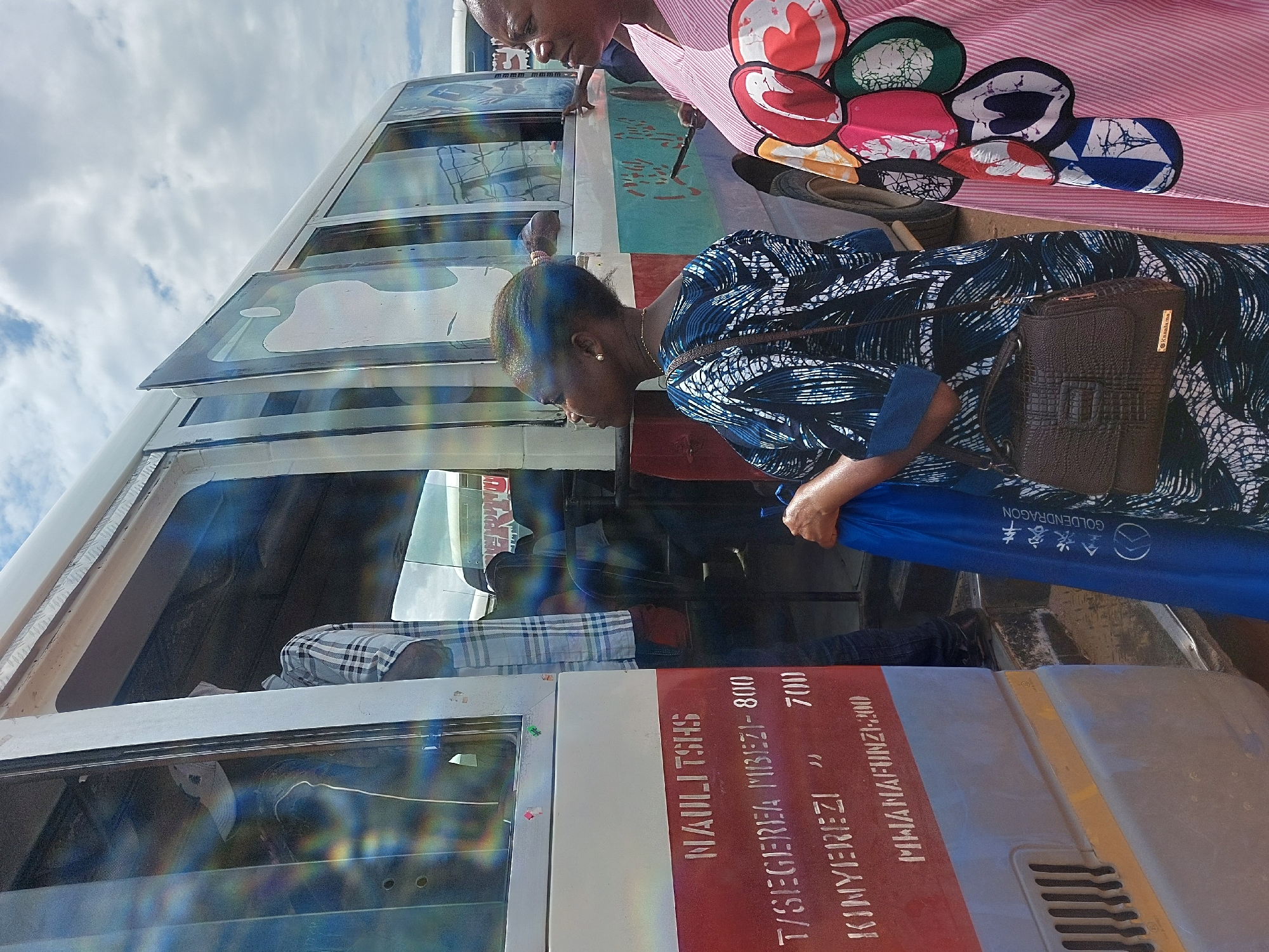 The image shows two women interacting with a bus. One woman, dressed in a blue and white patterned outfit with a brown purse, appears to be boarding the bus or placing an item inside. She is holding a blue bag with the "GOLDENDRAGON" logo. The other woman, wearing a pink striped shirt with colorful circular patterns, is standing next to the bus.
Text on the bus, "NAULITSHS T/SEGEREA MBEZI-800 KIMYEREZI MWANAFUNZI-200", indicates locations and possibly pricing, suggesting the bus is a public transport vehicle. The text mentions "Mbezi," a known area in Dar es-Salaam, Tanzania, indicating that this photo was taken in Dar es-Salaam.