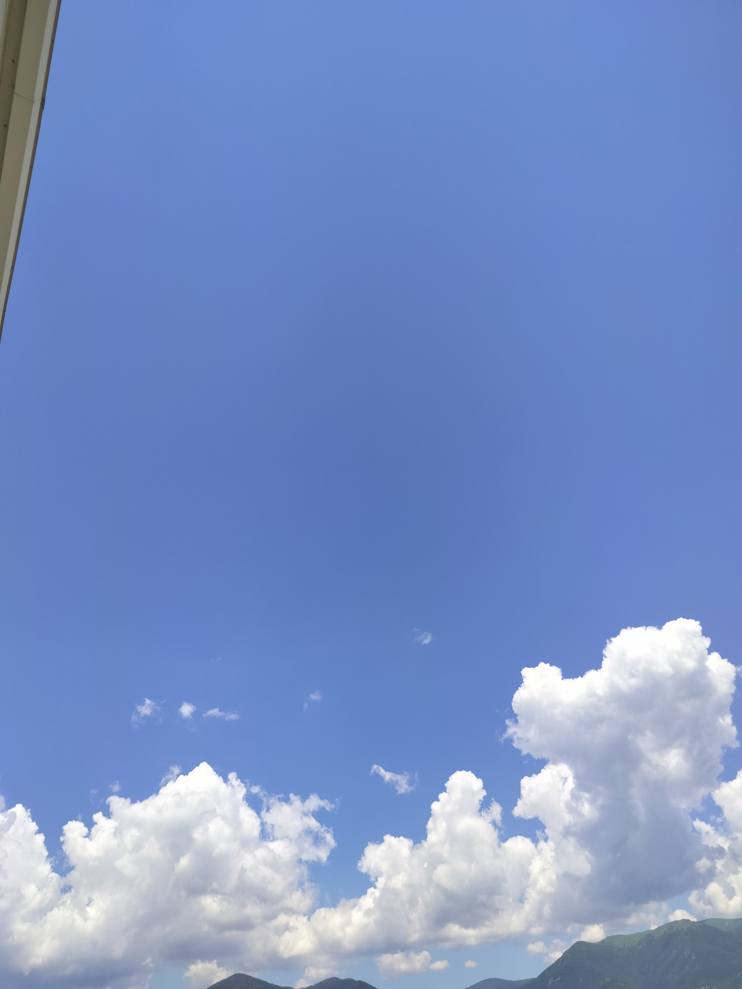 The image shows a clear blue sky with scattered white cumulus clouds. The bottom of the frame is dominated by a range of green, undulating mountains. The sky takes up the majority of the image, with the clouds concentrated towards the bottom. The perspective is from a low angle, looking upwards. There is a hint of a structure, possibly a building or awning, in the upper left corner of the image. The scene is serene and natural, characteristic of a sunny day in a mountainous region.