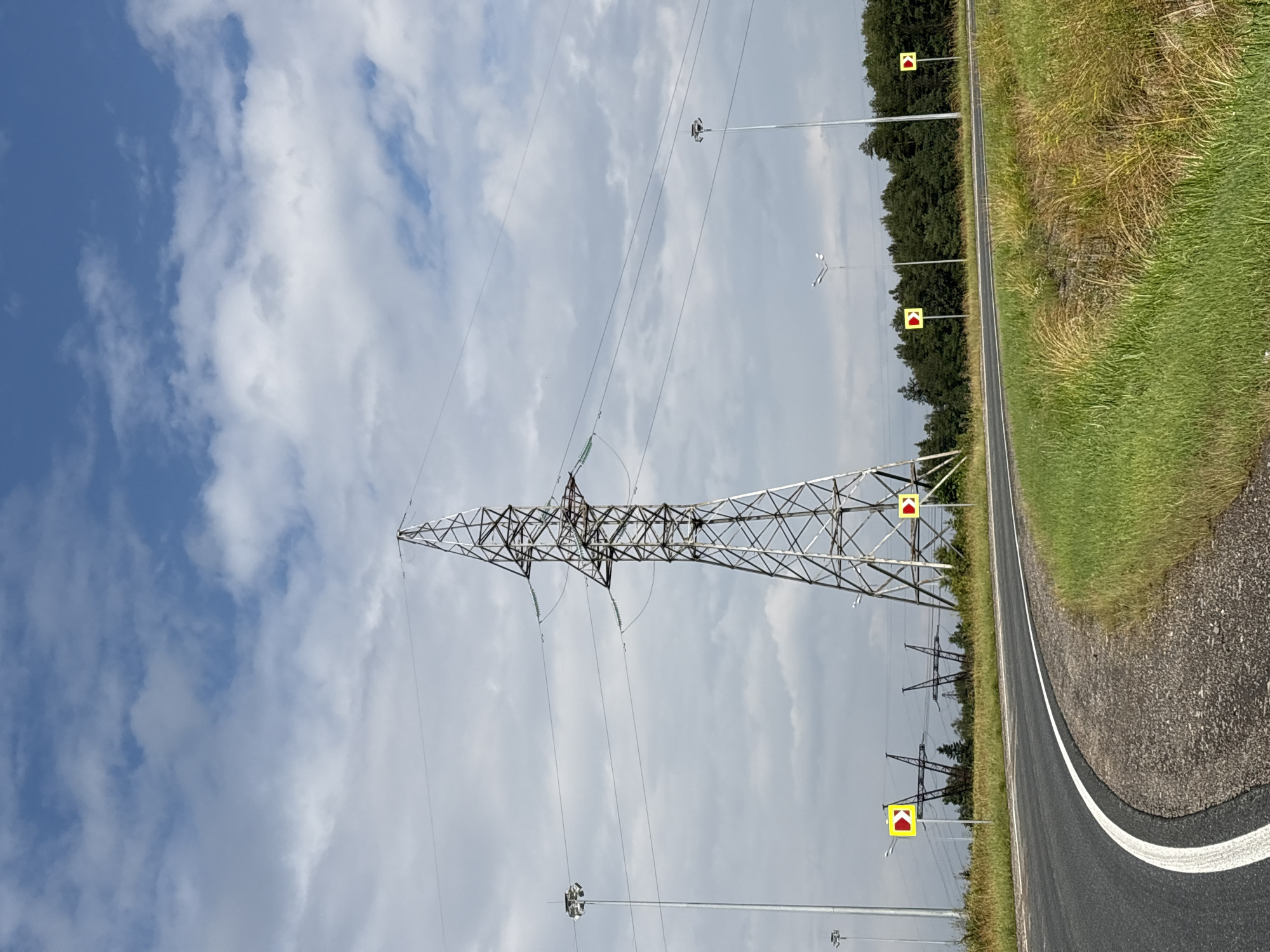 An asphalt road curves to the left, bordered by green grass. In the background, a large metal power line tower stands tall against a sky filled with white and gray clouds. Several smaller power line towers are visible further back, along with a line of trees. Two yellow road signs with red arrows indicating a turn are placed along the roadside. Tall streetlights are also present. The image appears to be taken on a cloudy day. The location is not specified in the image or the provided context, and therefore cannot be determined to be Perm, Russia.