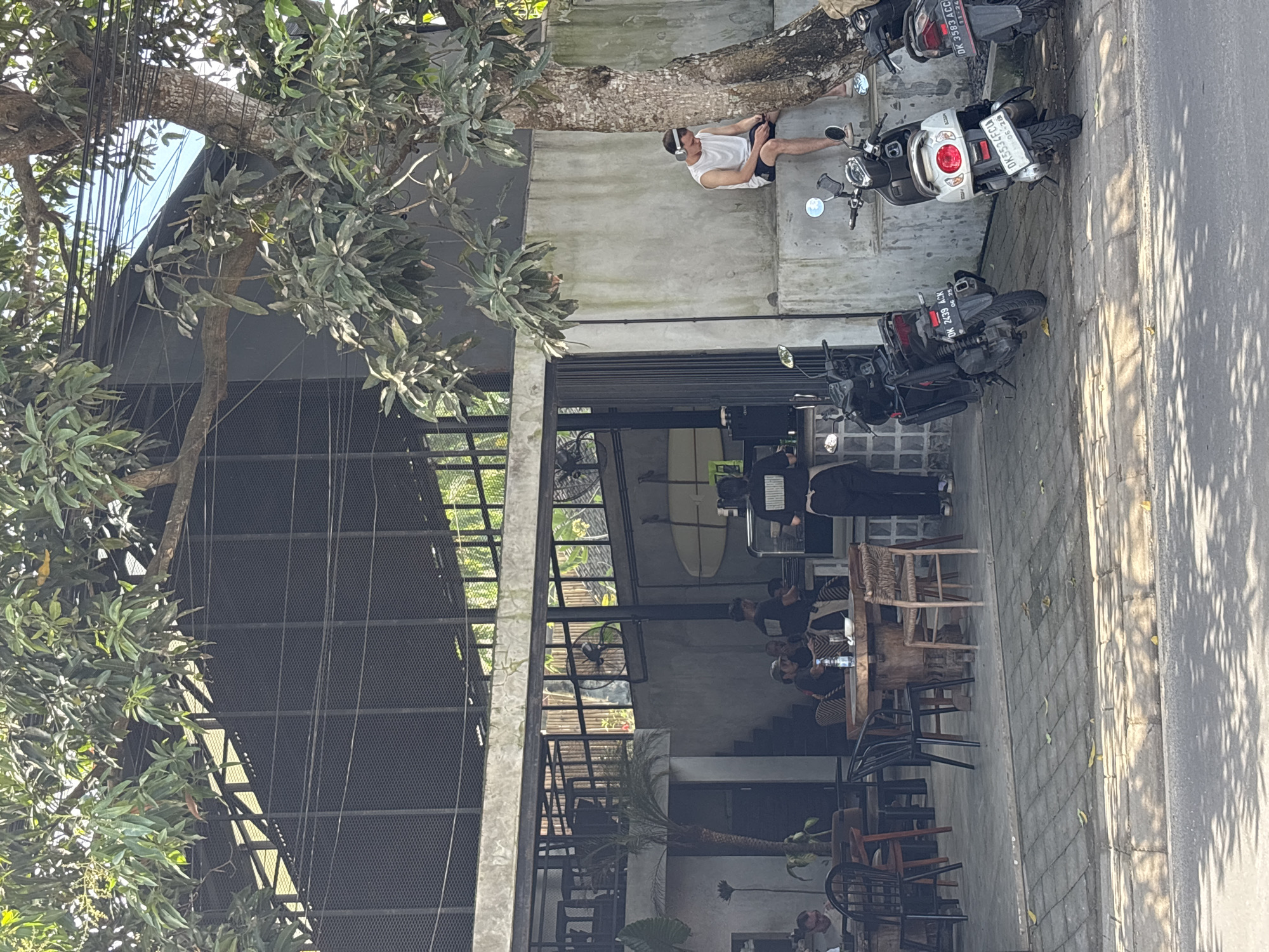 The image depicts the exterior of a two-story building with a partially open-air design. The location is Kuta Selatan, Indonesia, judging by the license plates on the motorbikes. The building features a grey concrete facade, with a large tree extending over the structure. The first floor is open, revealing tables and chairs occupied by several people, some of whom appear to be customers. There is also a surfboard visible inside. A staircase leads to the second floor, which is partially enclosed by a black mesh screen. Outside, on the pavement next to the building, are parked motorcycles. A man wearing headphones is sitting on the concrete ledge beside the tree and motorbikes. The street is paved and there are shadows cast by the trees.