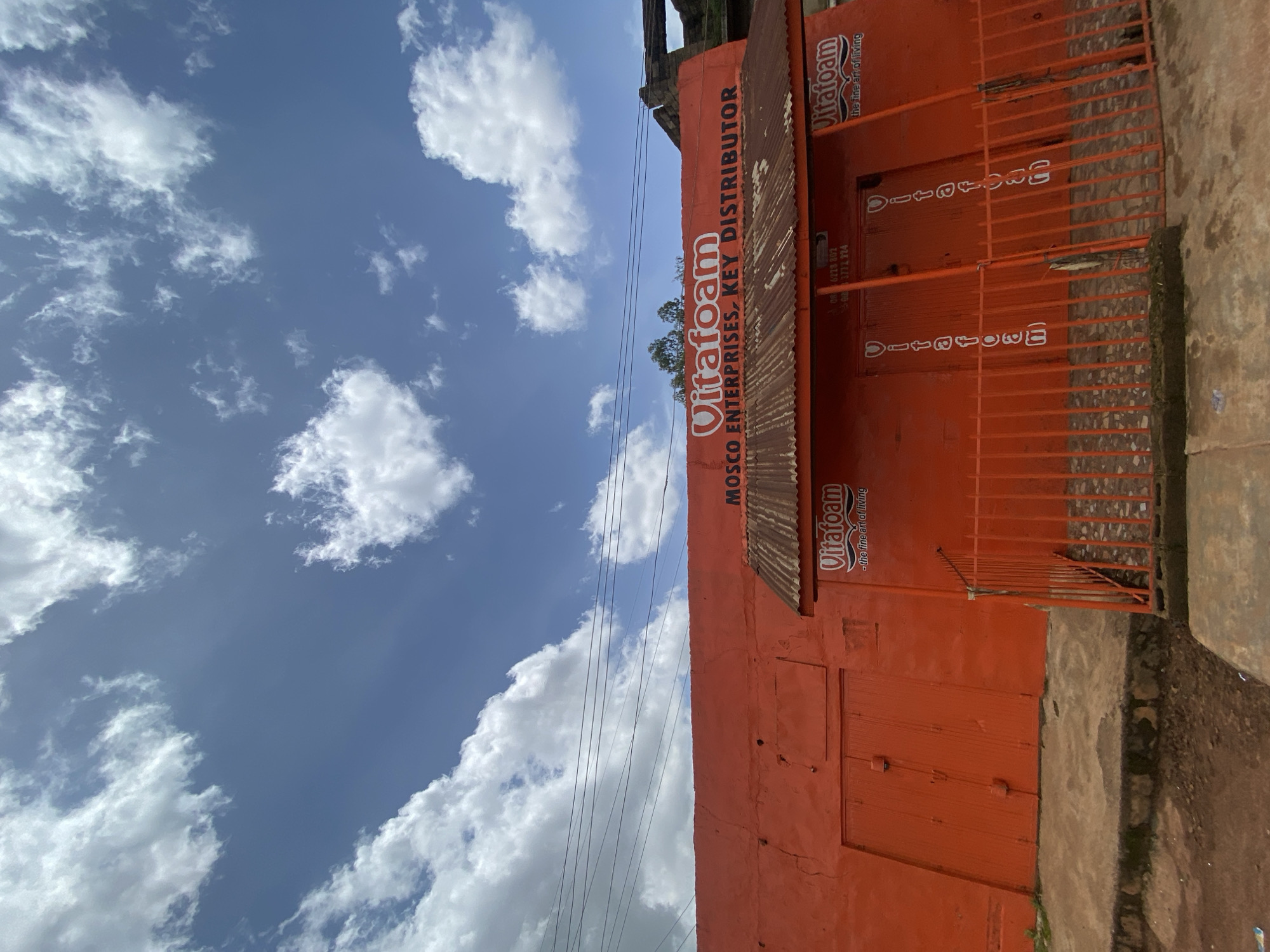 In Gura Top, Nigeria, a bright orange building stands under a blue sky with scattered white clouds. The building's facade is adorned with the white text "Vitafoam MOSCO ENTERPRISES, KEY DISTRIBUTOR." A rusted metal awning extends over the entrance area, where another "Vitafoam" logo is visible, accompanied by the slogan "-the fine art of living." Vertical "Vitafoam" logos are also present on the orange doors behind an orange metal fence. The ground in front of the building is a mixture of concrete and dirt. Overhead, power lines crisscross the sky.
