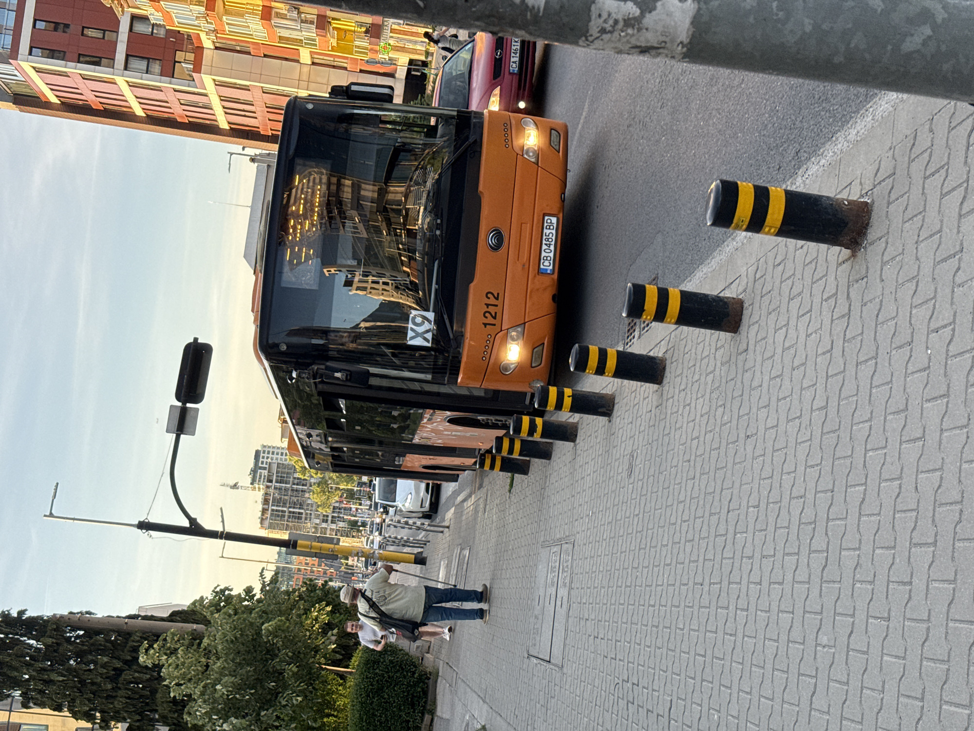 The image shows a street scene in Sofia, Bulgaria. An orange public bus with license plate "CB 0485 BP" and the number "1212" is driving on the right side of the road. A red Opel car with license plate "CA 1461 K" can be seen behind the bus. A sidewalk with gray pavement is on the right. On the sidewalk, there are black metal bollards with yellow stripes. Two people are walking along the sidewalk. One person appears to be taking a photo. The background shows buildings and a blue sky. A traffic light is also visible on the left.