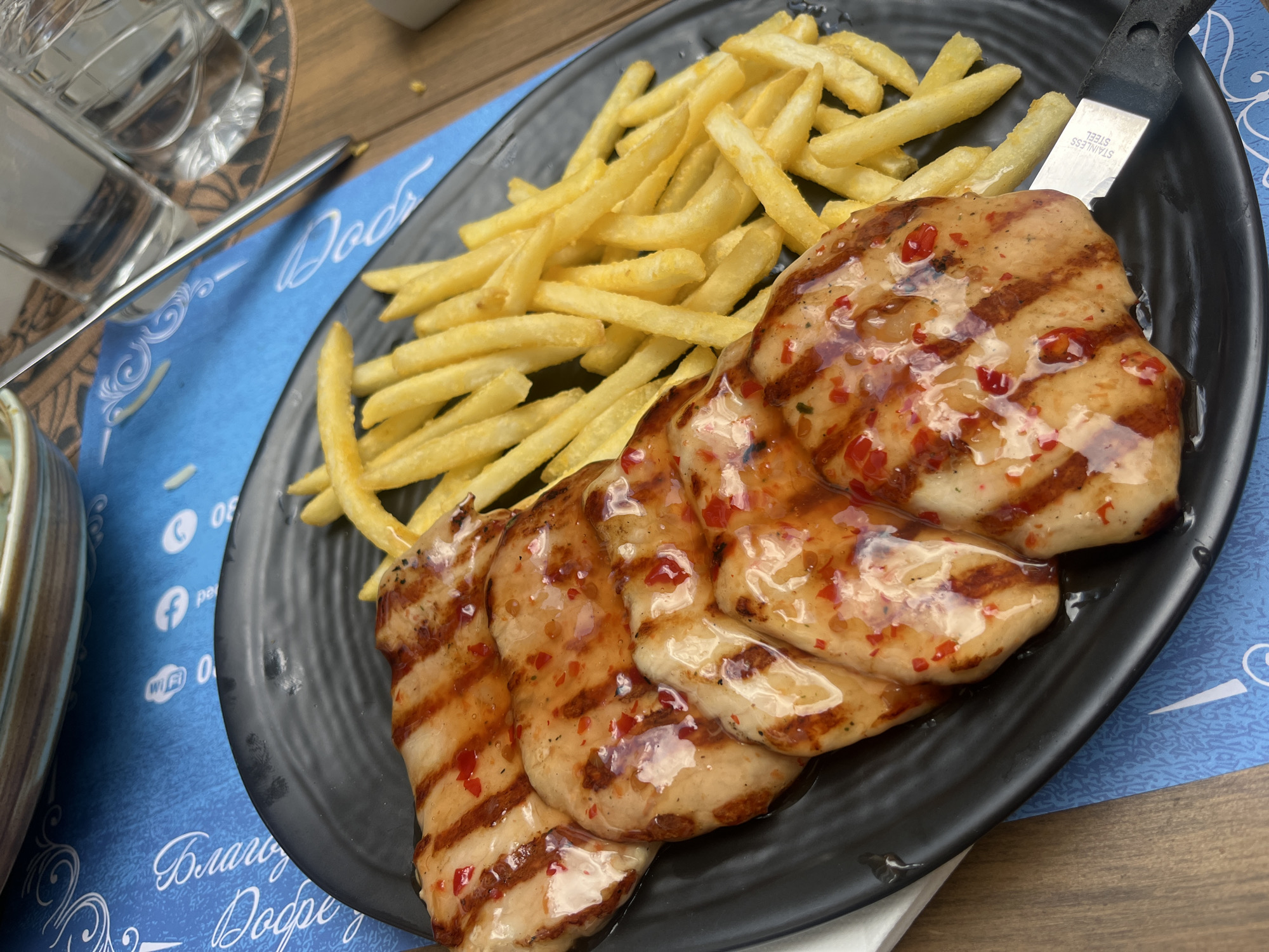 A close-up shot shows a black plate of grilled chicken with chili glaze and french fries. A steak knife lies across one of the chicken pieces. The plate rests on a blue placemat with white decorative patterns and social media icons, including a Facebook icon and a Wi-Fi symbol. Bulgarian Cyrillic text is visible on the placemat. The scene appears to be set outdoors at a restaurant in Stara Zagora, Bulgaria.