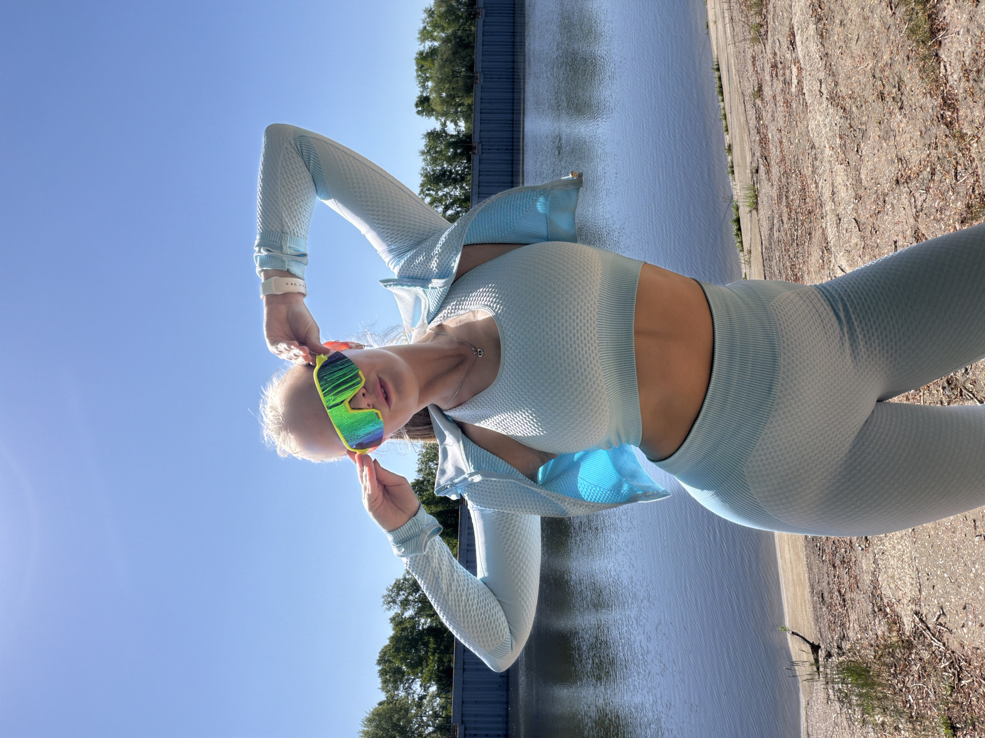 The image shows a woman in athletic wear standing outdoors. She is wearing a light blue matching set consisting of leggings, a sports bra, and a long-sleeved jacket. She is also wearing multicolored sunglasses. She is standing on a dirt surface near a body of water, possibly a river. Behind her, there is a concrete or metal structure. The sky is clear and blue. The location is the city of Chaikovsky, Russia.