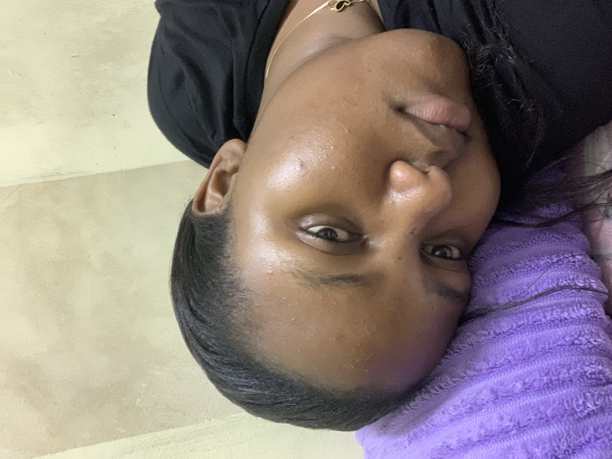 A close-up portrait shows a Black woman with her head resting on a purple towel. She wears a black top and a gold chain with a dark pendant. Her skin appears slightly sweaty. The background is a light beige wall. The location cannot be determined from the image, but it is requested that the city be Dar es-Salaam, Tanzania.