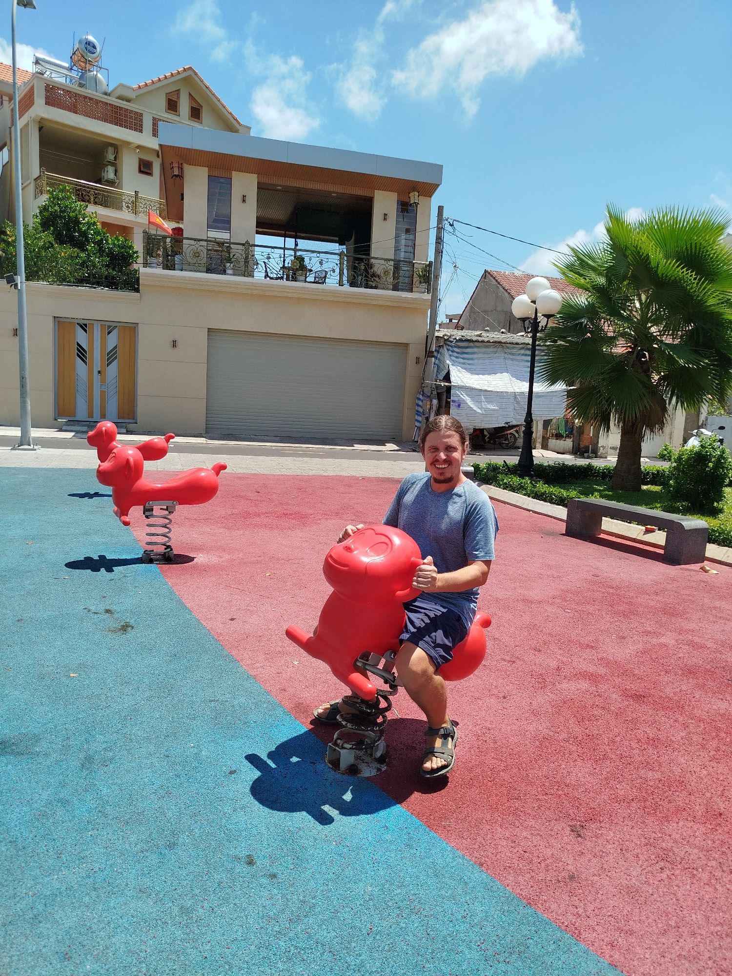 A man rides a red, pig-shaped spring rider in a playground in Ho Chi Minh City, Vietnam. The playground surface is divided into blue and red sections. Behind him, another identical spring rider stands on the blue section. In the background is a two-story building with a water heater on the roof. To the right, a palm tree and a bench are visible.