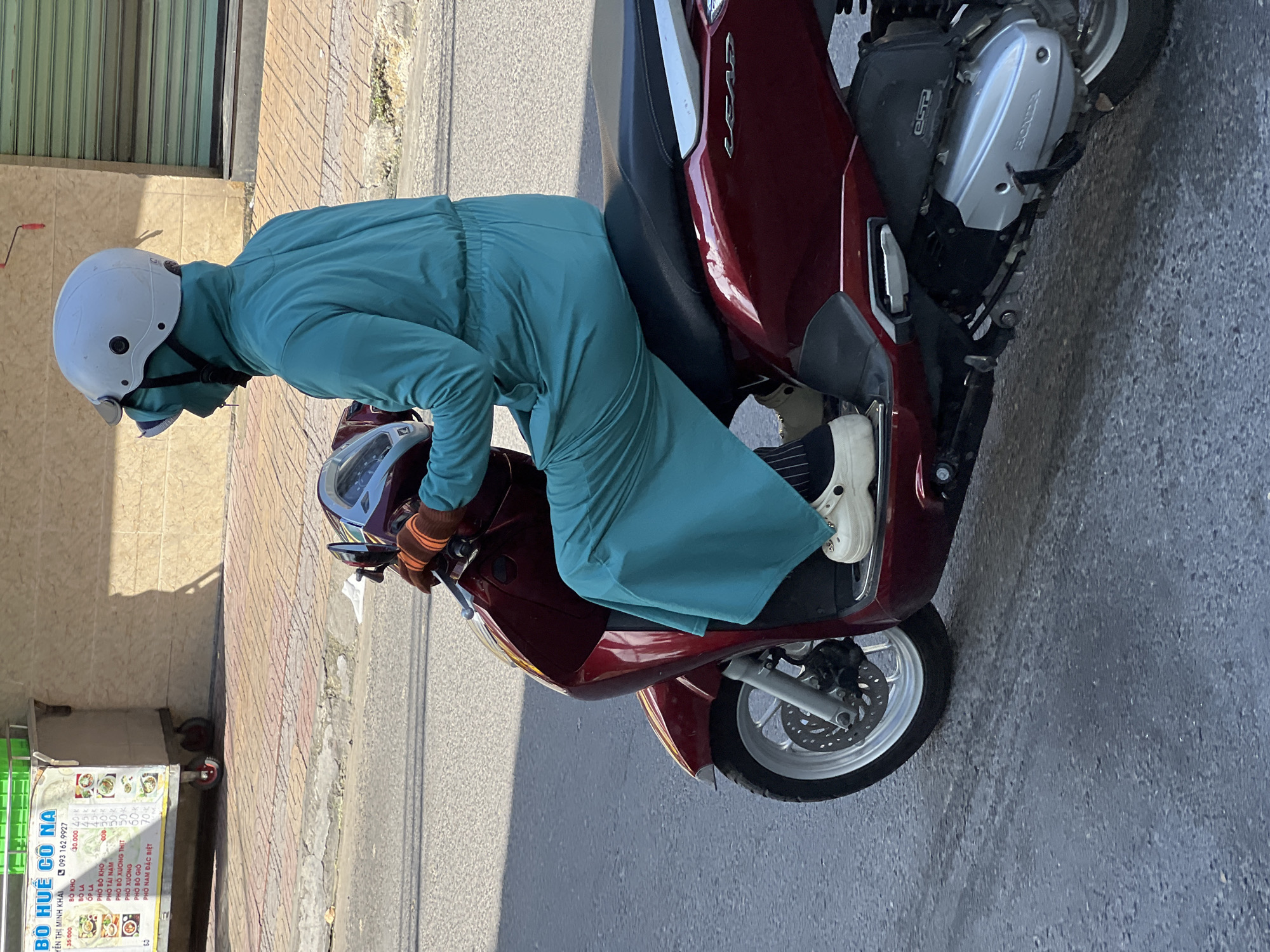 In Nha Trang, Vietnam, a person is riding a maroon Honda Lead scooter. They are wearing a full-coverage teal outfit, a white helmet with a visor, and a blue face mask. The person is also wearing tan Crocs with black socks. The scooter has a dark brown seat and silver accents. In the background, a food cart with a menu is visible, displaying prices in Vietnamese Dong and offering items like "Pho Bo Kho" and "Pho Bo Gio." The street has a tiled sidewalk and asphalt road, with a light-colored building and a green metal shuttered storefront in the background. The sun is casting shadows on the ground.