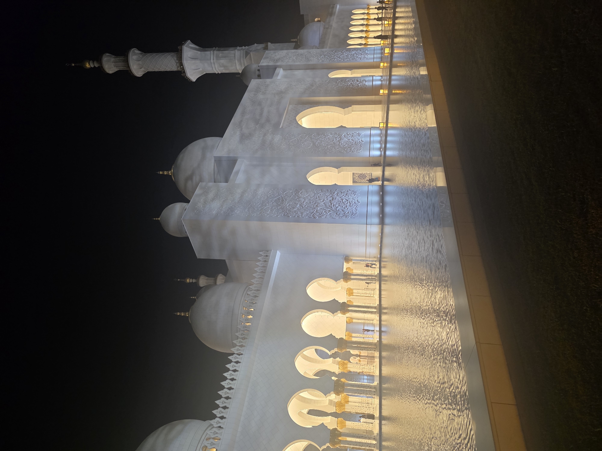 The photograph captures the Sheikh Zayed Grand Mosque in Abu Dhabi, United Arab Emirates, at night. The mosque's white marble facade is illuminated by a soft, warm light, highlighting its intricate architectural details. The building features multiple domes and minarets, with a prominent tall minaret on the right side of the frame. A reflective pool of water in the foreground mirrors the illuminated structure, creating a symmetrical and tranquil atmosphere. Several figures, appearing as silhouettes, are visible walking along the colonnades and near the water's edge, suggesting visitors or staff. The sky above is dark, indicating nighttime, and a subtle haze or mist might be present, softening the overall illumination. The surrounding area appears to be a manicured lawn.