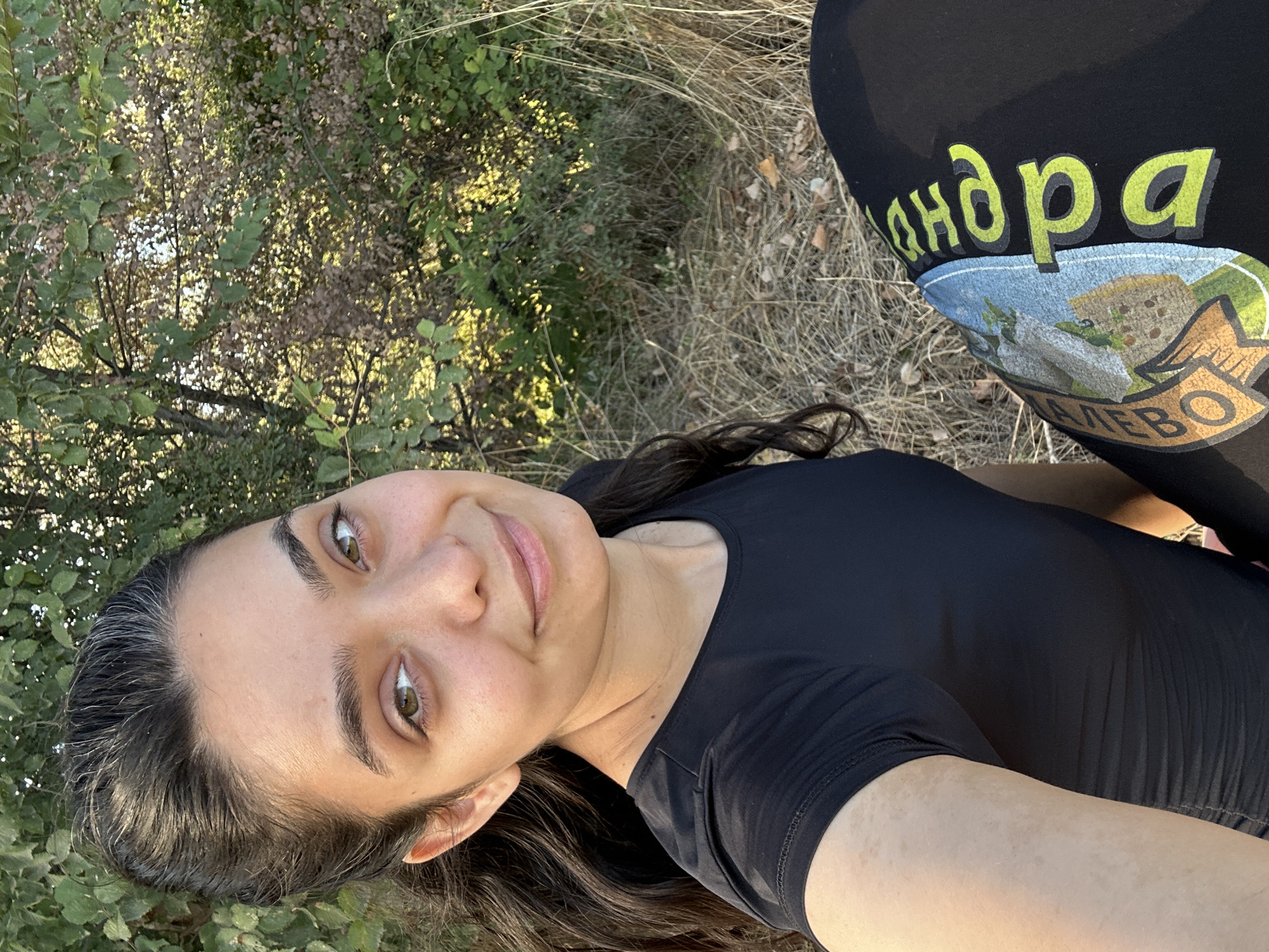 A young woman with dark hair pulled back and a few gray strands visible is taking a selfie outdoors. She is wearing a black t-shirt and black leggings with a yellow and green design on them. The background consists of dry grass, trees, and foliage, suggesting a natural or park-like setting. The lighting indicates it is daytime, possibly late afternoon due to the warm tones. The image does not contain information to determine a specific location within the city of Malevo, Bulgaria.