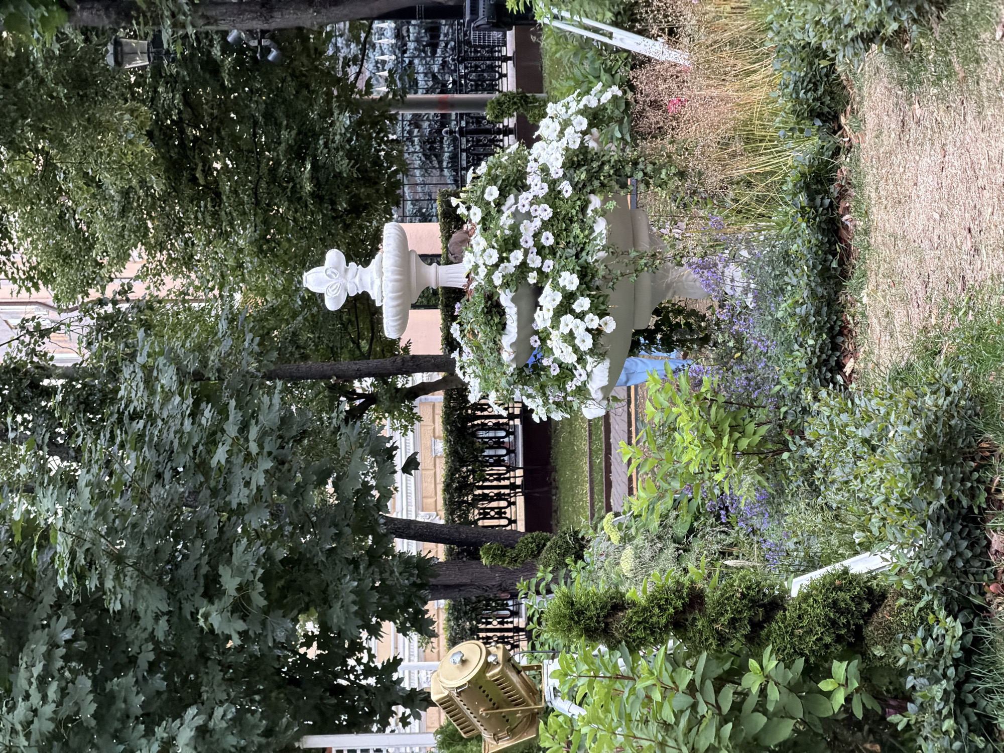 A tiered white fountain serves as a centerpiece in a lush garden, overflowing with white petunias. To the left, a golden spotlight is positioned on a tripod amidst green foliage. A winding dirt path leads through the garden, which is bordered by manicured topiaries, flowering plants, and various shrubs. In the background, trees and a building with light-colored walls and dark trim are visible, suggesting an urban park setting. The scene is likely located in a public garden or park within the city of Moscow, Russia, given the architectural style of the building and the ornamental garden design.