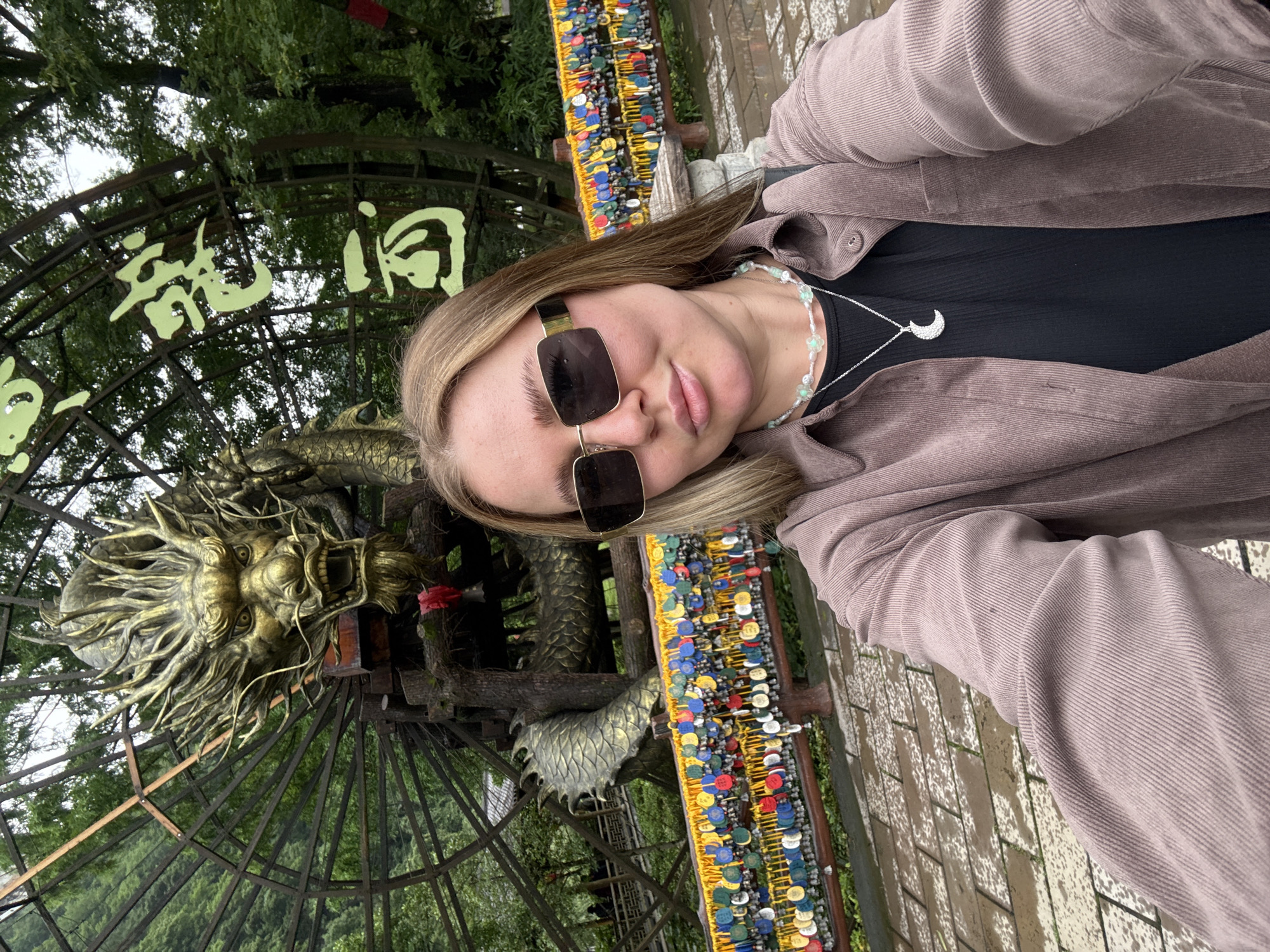 A selfie taken by a person with shoulder-length blonde hair and sunglasses. The person is wearing a black top and a light brown corduroy jacket, adorned with a white floral necklace with a silver moon pendant. In the background, there's a large golden dragon sculpture within a dark metal frame, with Chinese characters visible above it. The frame and nearby railings are adorned with numerous colorful medallions. The ground appears to be tiled. This image appears to be taken in Jundiping, China.